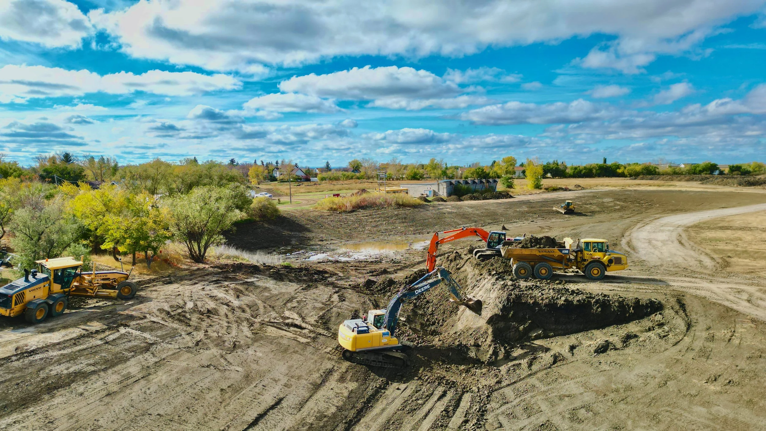 Construction site with excavators and dump trucks on dirt ground, trees on the left and a blue sky with clouds overhead.