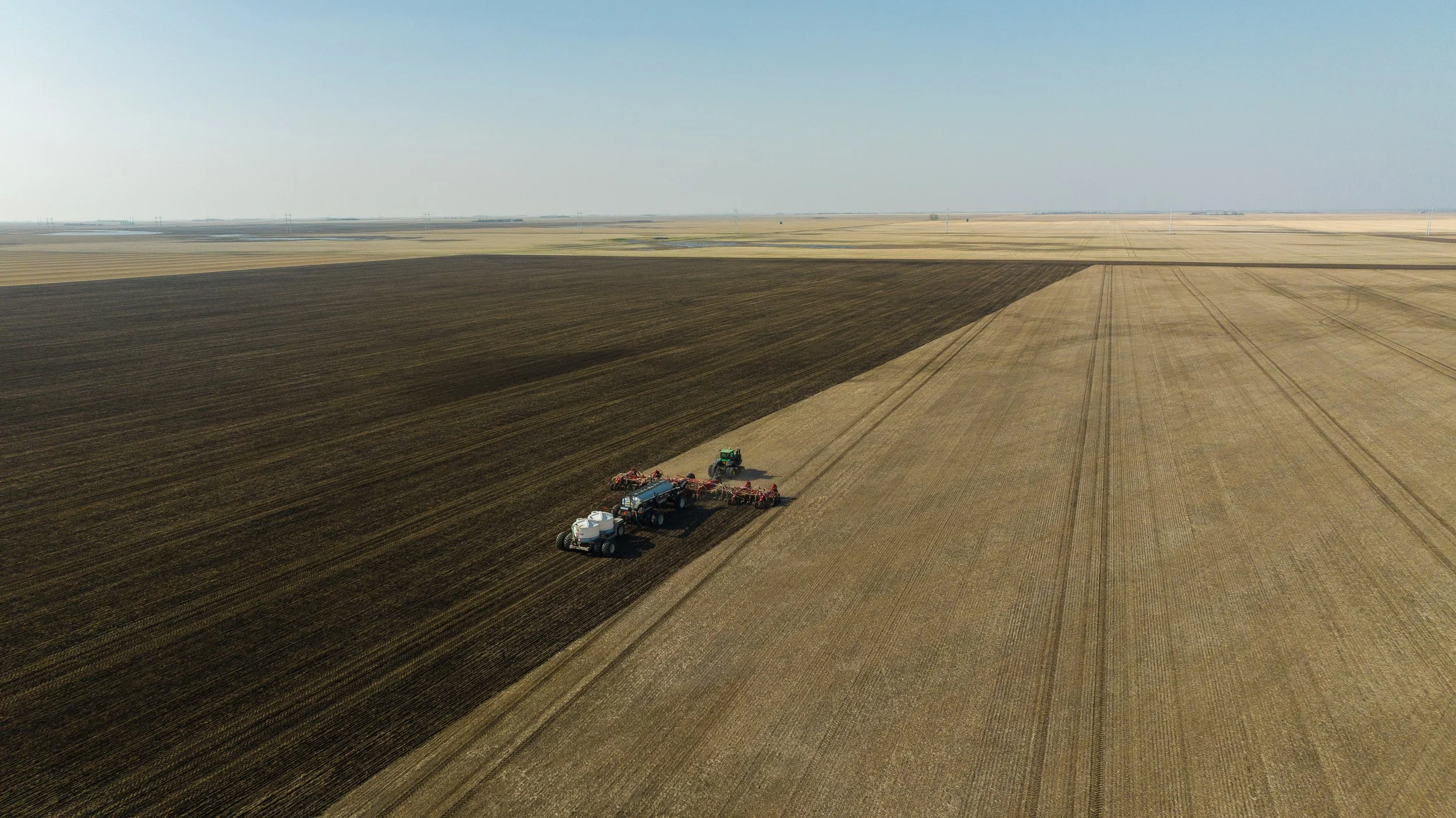 Agricultural machinery working on large fields of dry and dark soil, with distant power lines and a flat horizon under a clear sky.