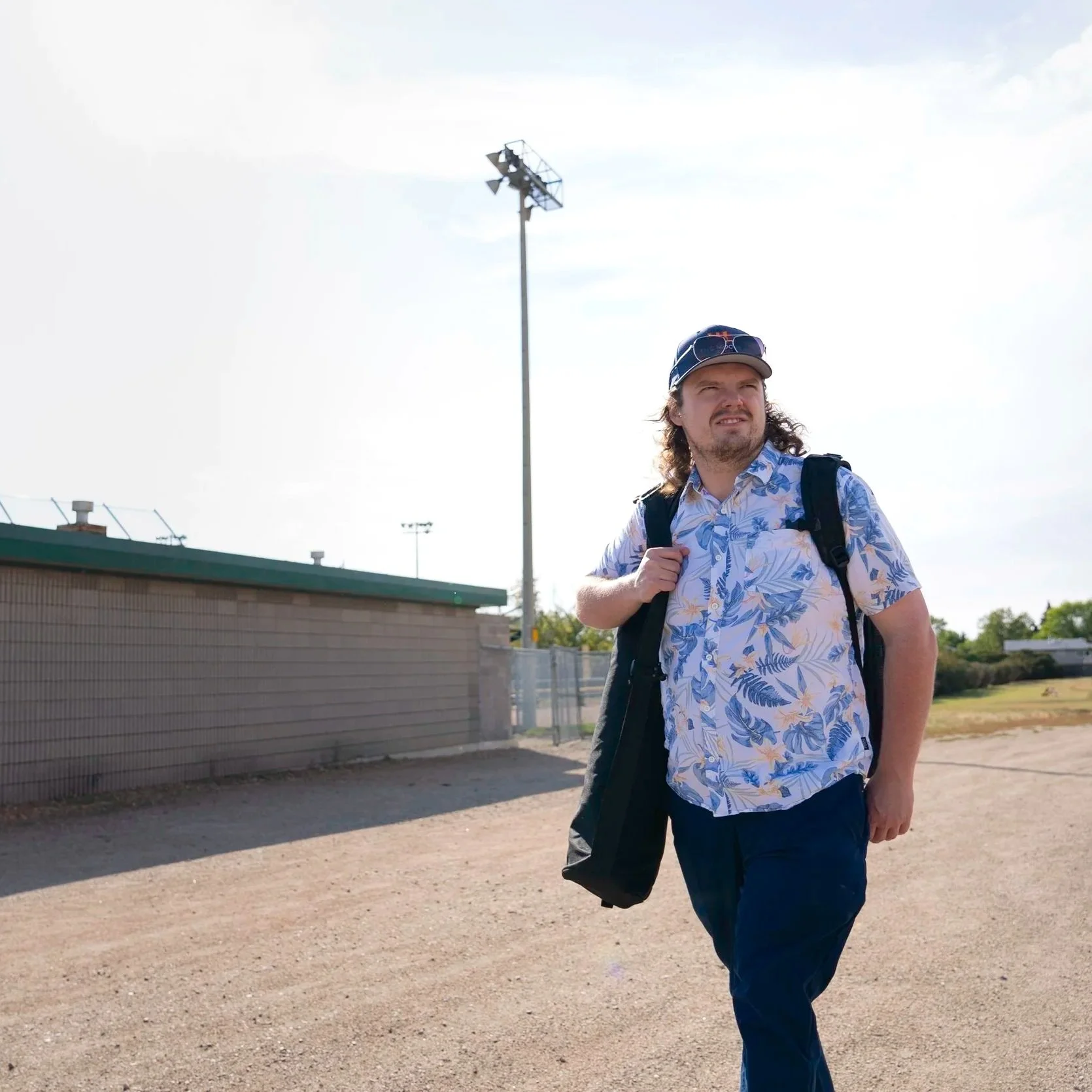 A man with long hair and a beard, wearing a blue floral shirt and sunglasses, walking outdoors with a backpack, on a clear day with a stadium light in the background.