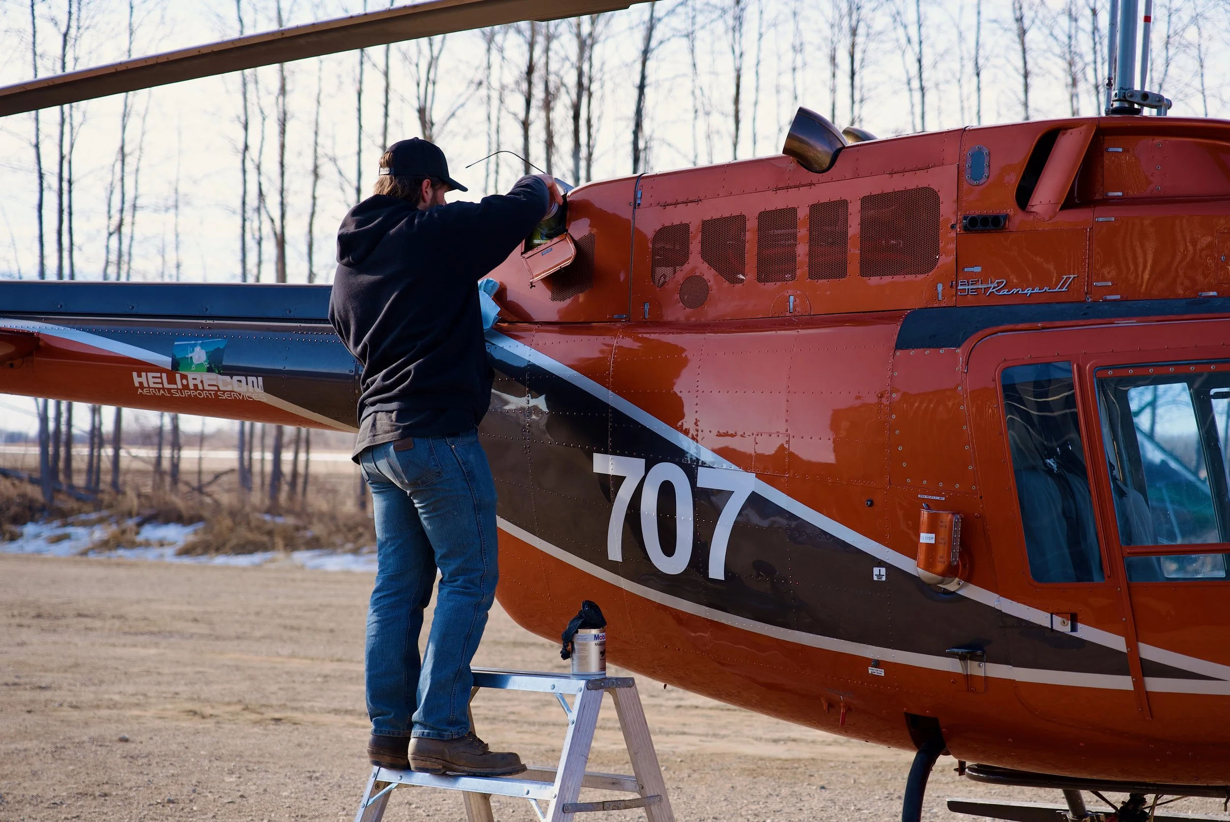 A man standing on a step ladder working on a red helicopter outdoors, with trees and a partly cloudy sky in the background.