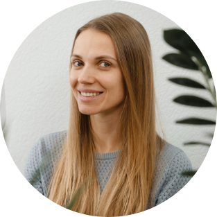 A woman with long blonde hair smiling, standing indoors near a white wall and a leafy plant.