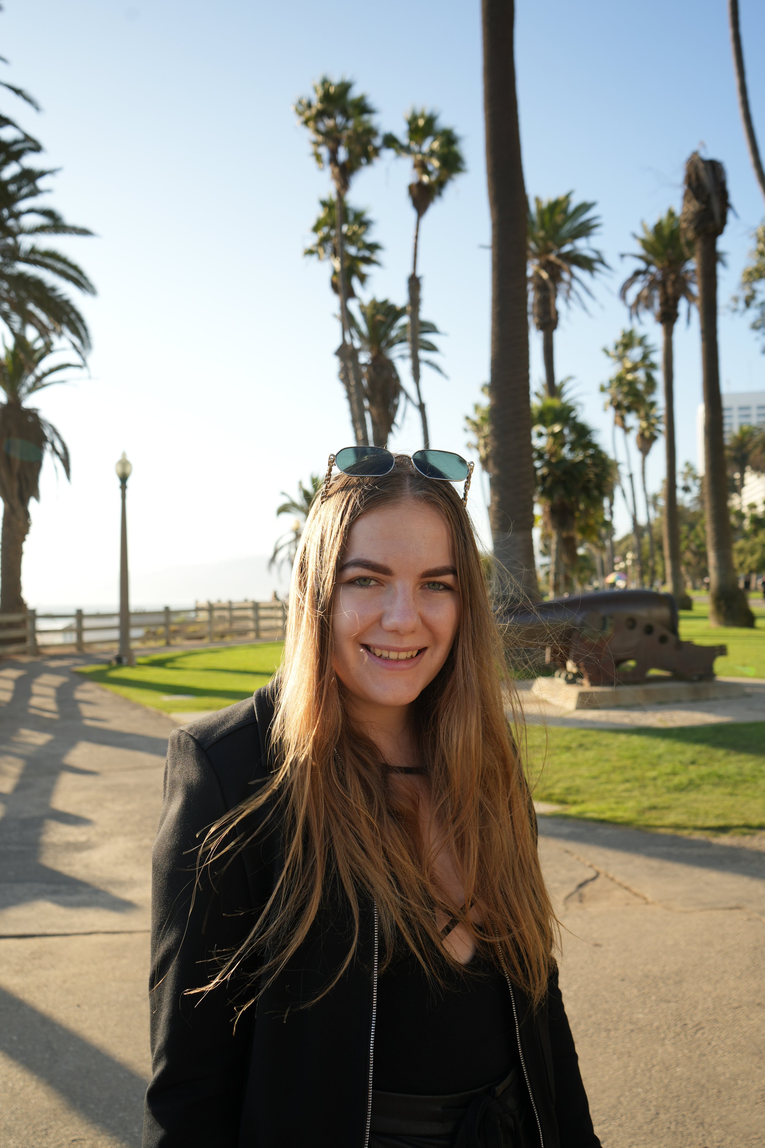 A young woman with long hair wearing sunglasses on her head, smiling at the camera, standing outdoors in front of palm trees on a sunny day.