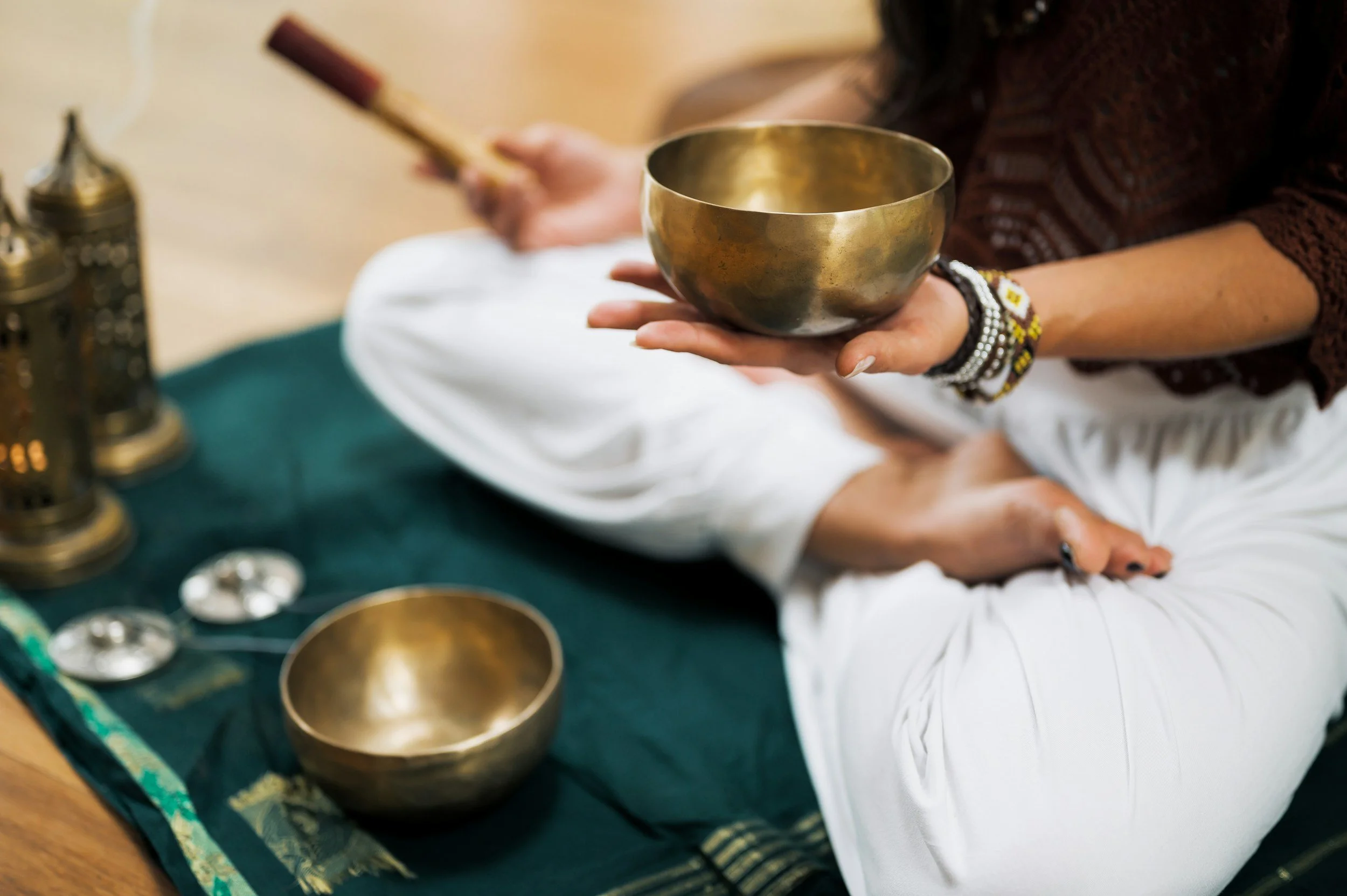 Person sitting cross-legged on a green cloth with brass singing bowls and decorative items, holding a brass singing bowl in hand, wearing white pants and multiple bracelets.