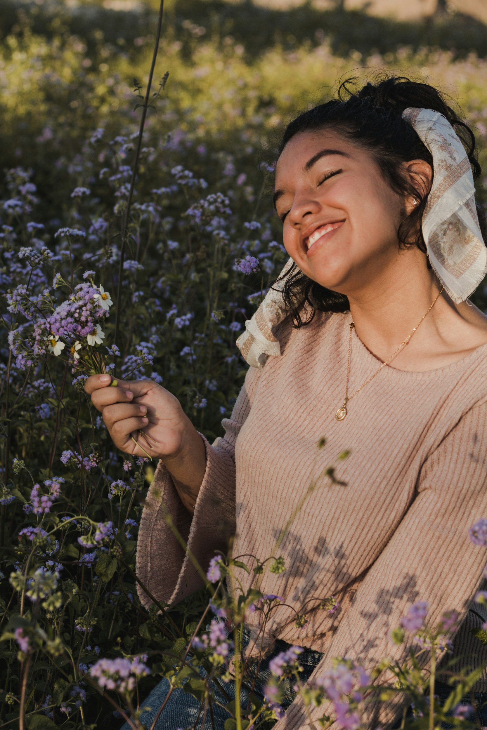 A young woman smiling with eyes closed, holding a small bouquet of flowers, standing in a field of purple flowers during sunset.