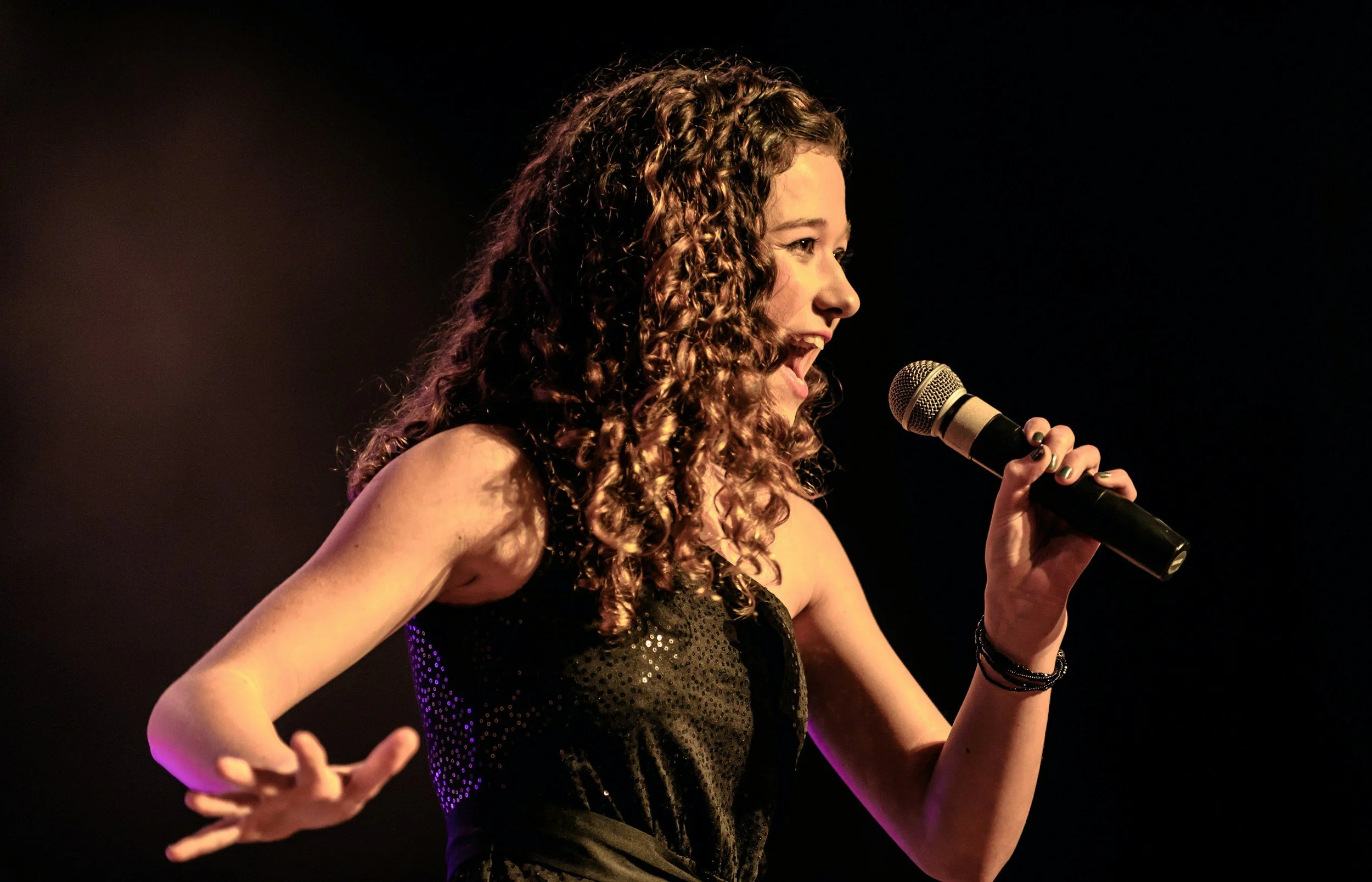 Young woman with curly hair singing into a microphone on stage, wearing a black dress with sequins.