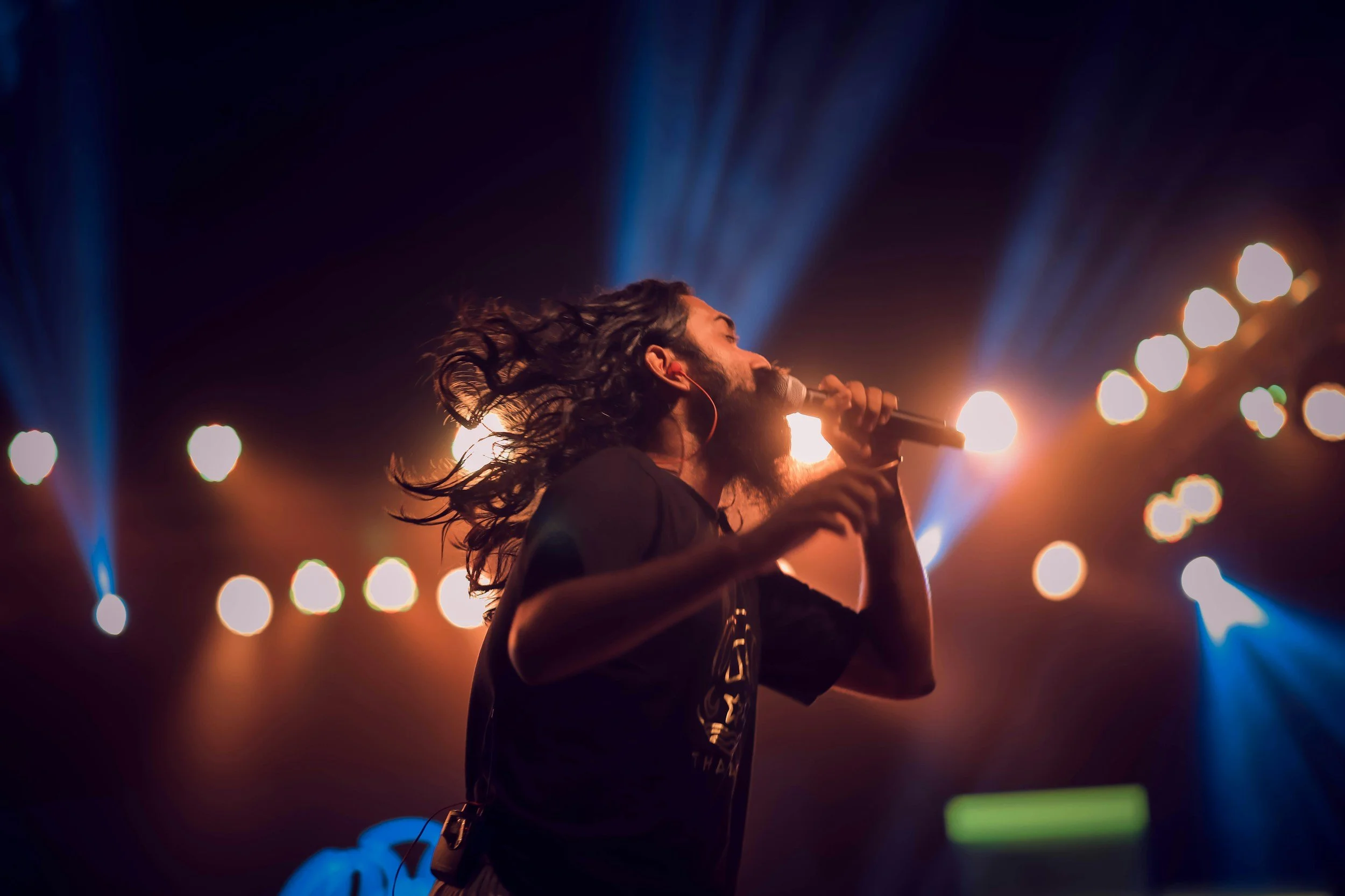 A male singer with long, curly hair and beard performing on stage with colorful stage lights shining behind him.