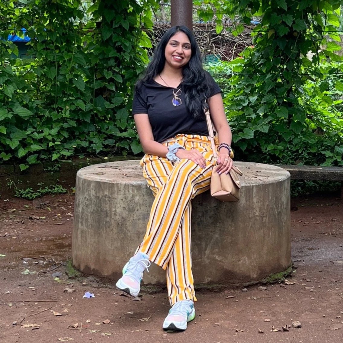 A young woman with long black hair sitting on a large concrete sculpture in a park, surrounded by green foliage, smiling at the camera. She is wearing a black t-shirt, yellow striped pants, colorful sneakers, and carrying a beige handbag.