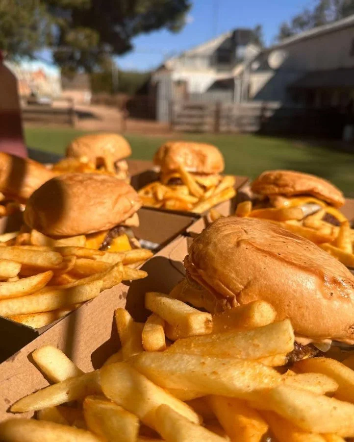 Close-up of burgers with French fries in cardboard containers, outdoor setting with trees and houses in the background.