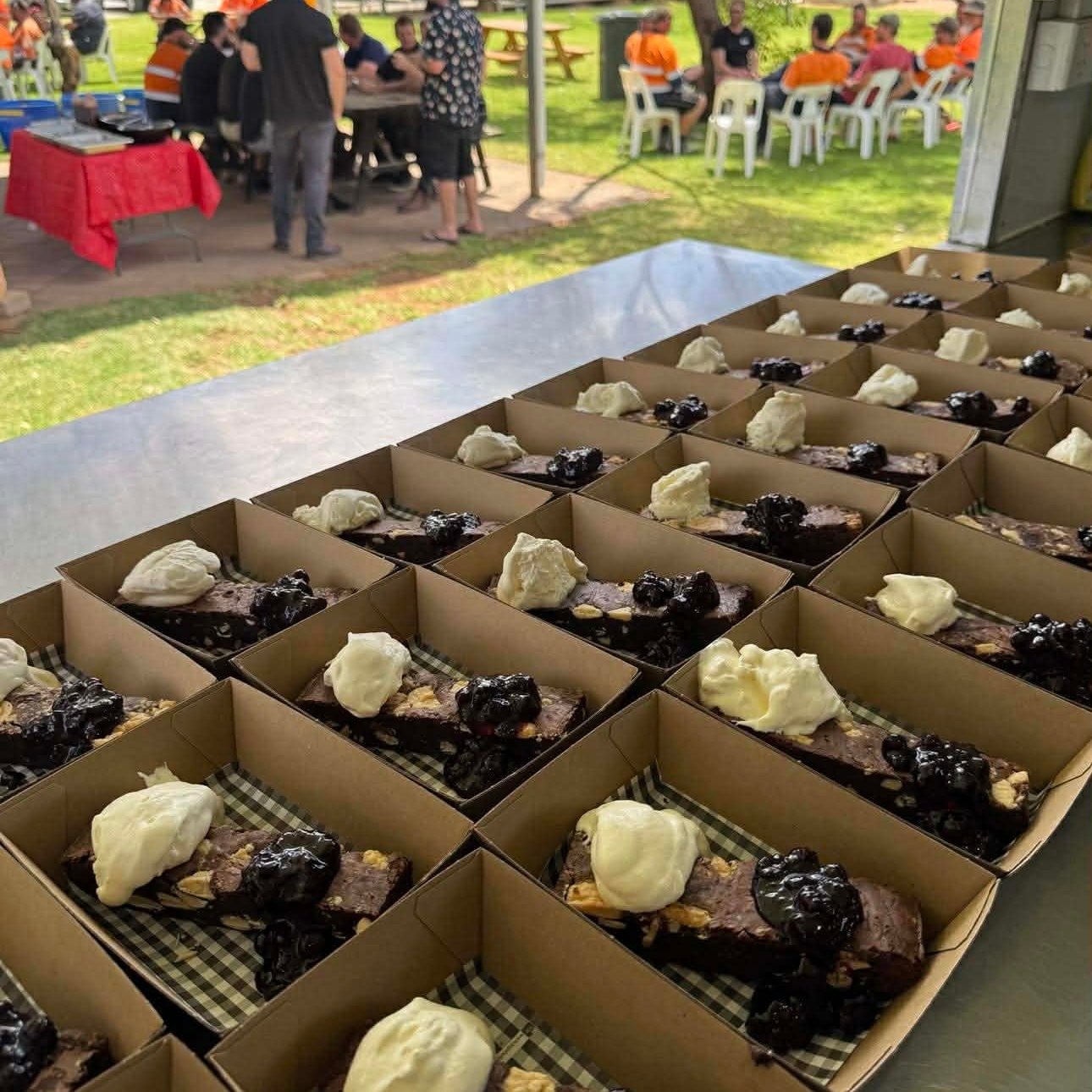 Rows of small cardboard trays filled with slices of beef topped with dollops of whipped cream and blueberries, set on a table at a gathering outdoors with people sitting at tables in the background.