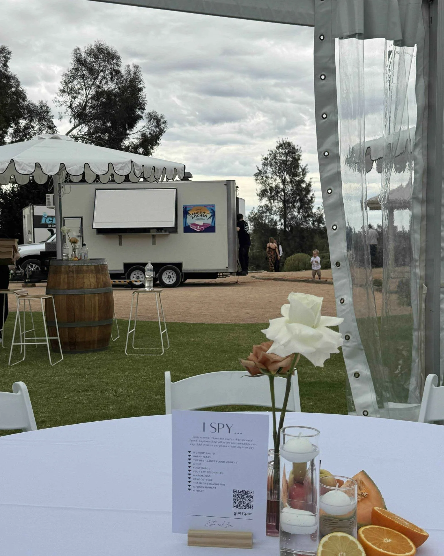 A table with a white tablecloth, flowers, candles, and sliced oranges and lemons, with a menu card titled 'I SPY...' on it, outdoor event with a food truck, trees, cloudy sky, and a few people in the background.