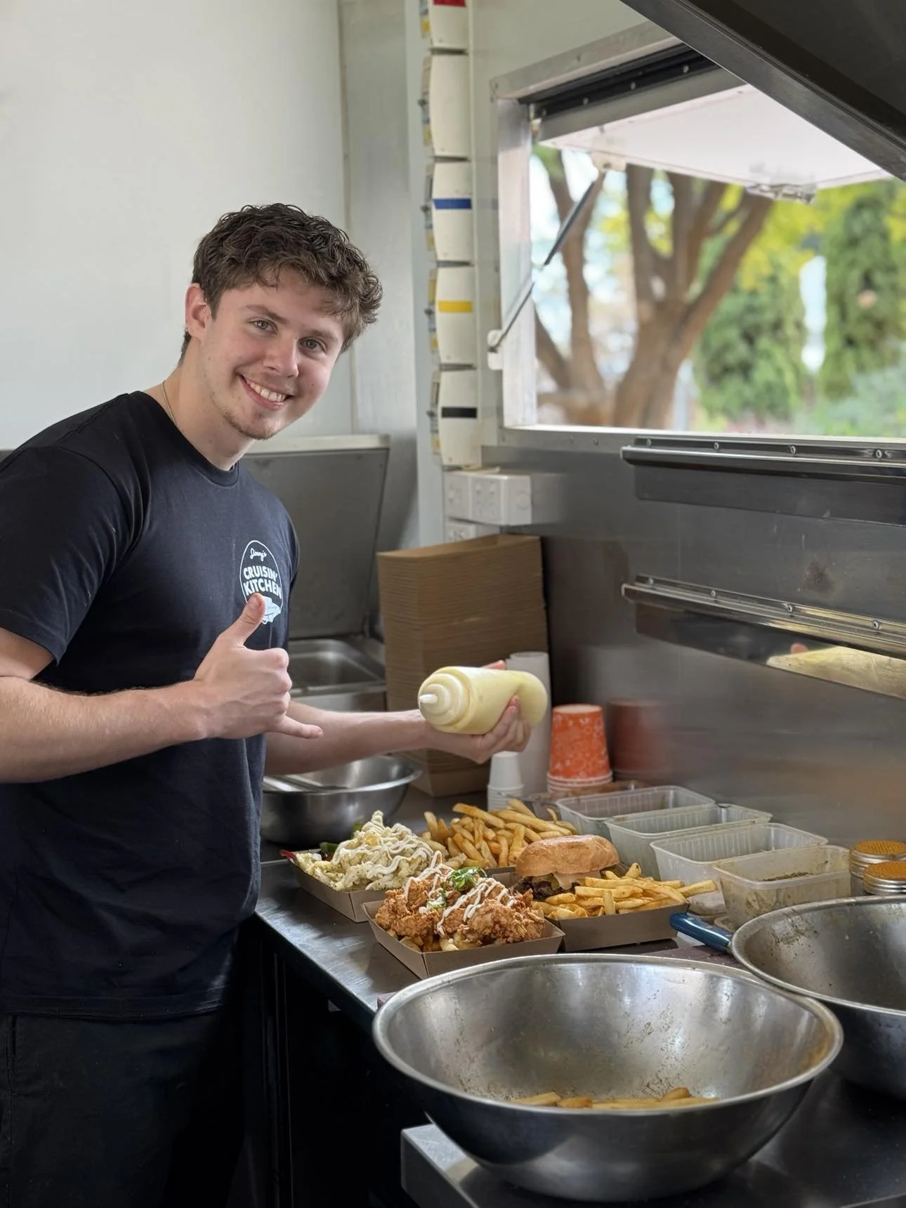 Young man in a black t-shirt preparing food in a food truck, holding a squeeze bottle, with various food containers and trays of fries and fried chicken on the counter.