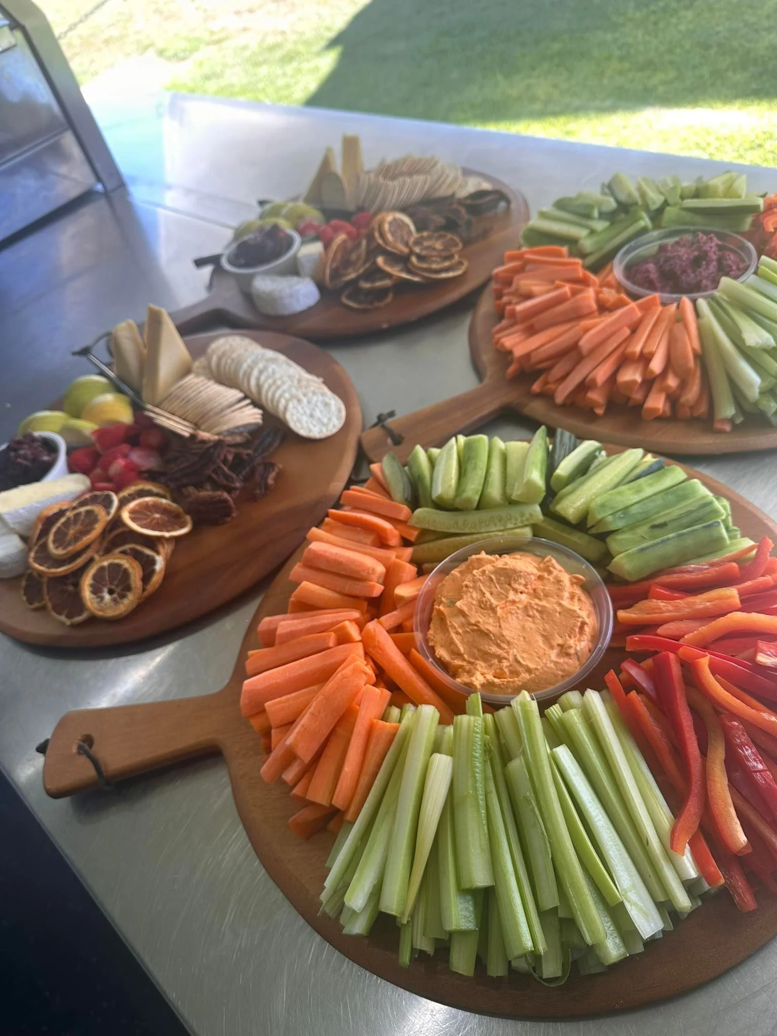 Three wooden charcuterie boards with vegetables, cheeses, dried fruits, and dips on a countertop with a grassy outdoor background.