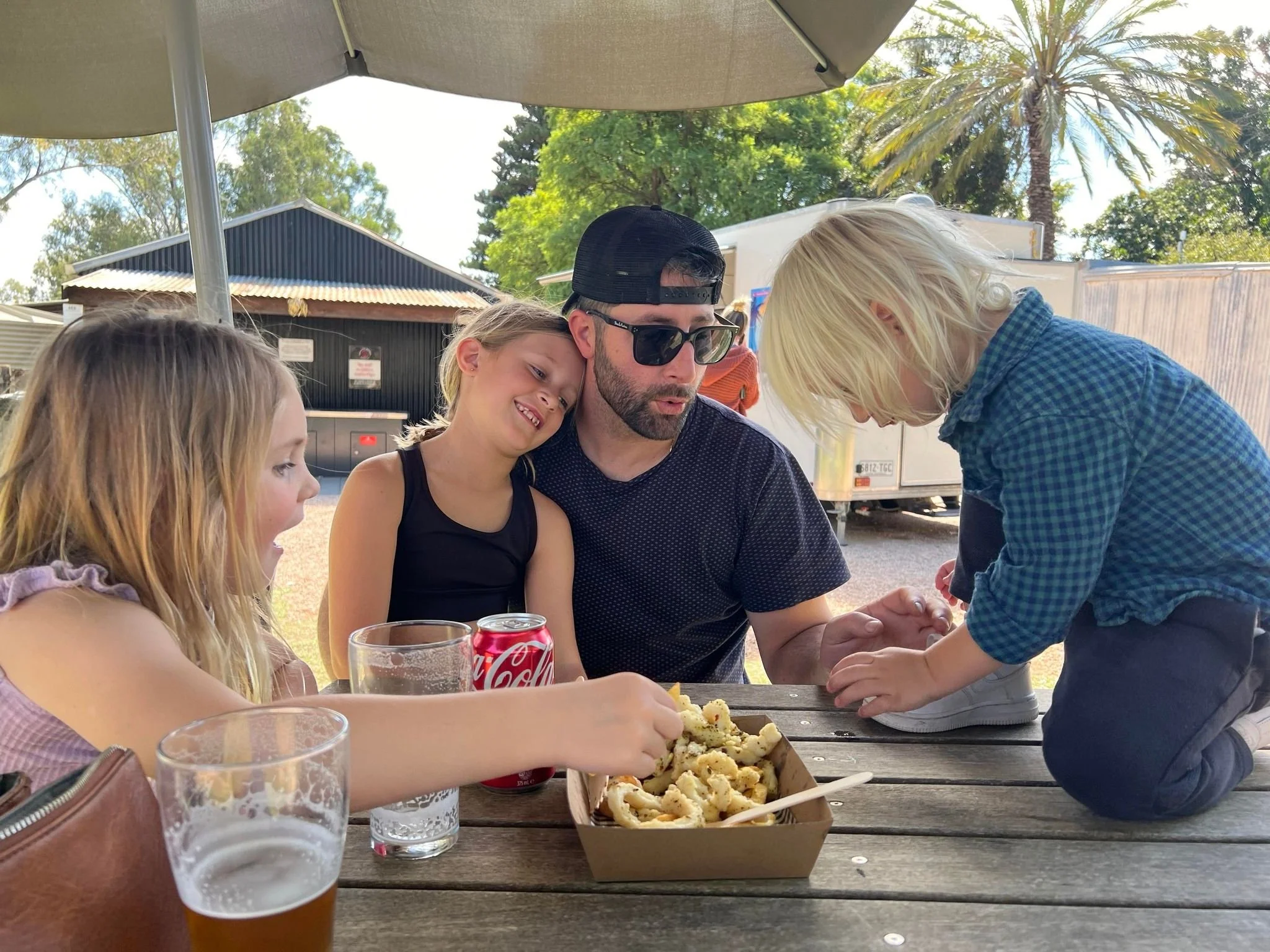 A family of five sitting at a picnic table outdoors. A man with sunglasses and a black cap is holding a small child on his lap. The child is leaning in close to him. Two young girls are sitting beside the man, one reaching for food. A bowl of curly f