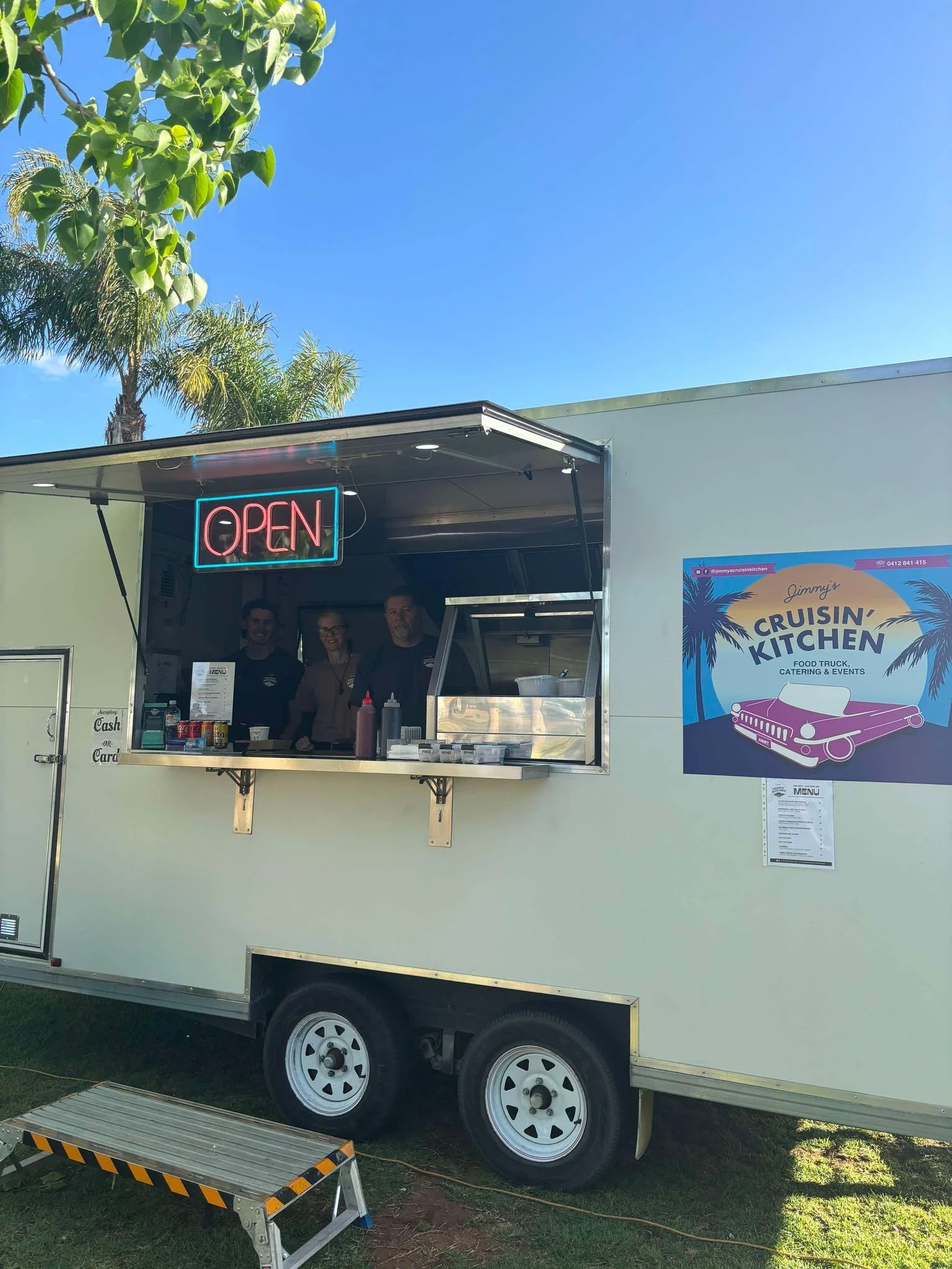 Food truck with a neon open sign, three staff members inside, a sign on the side reads 'Jimmy's Cruisin' Kitchen,' palm trees and a blue sky in the background.