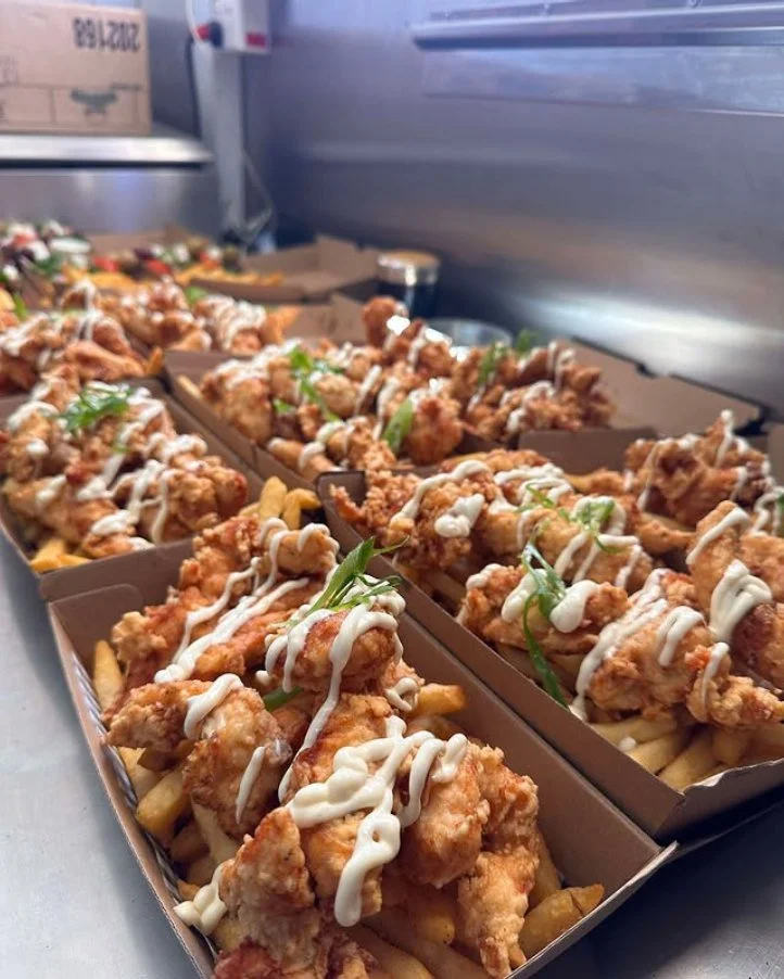 Boxes of fried chicken tenders topped with white sauce and green garnish, served over fries in a food kitchen setting.
