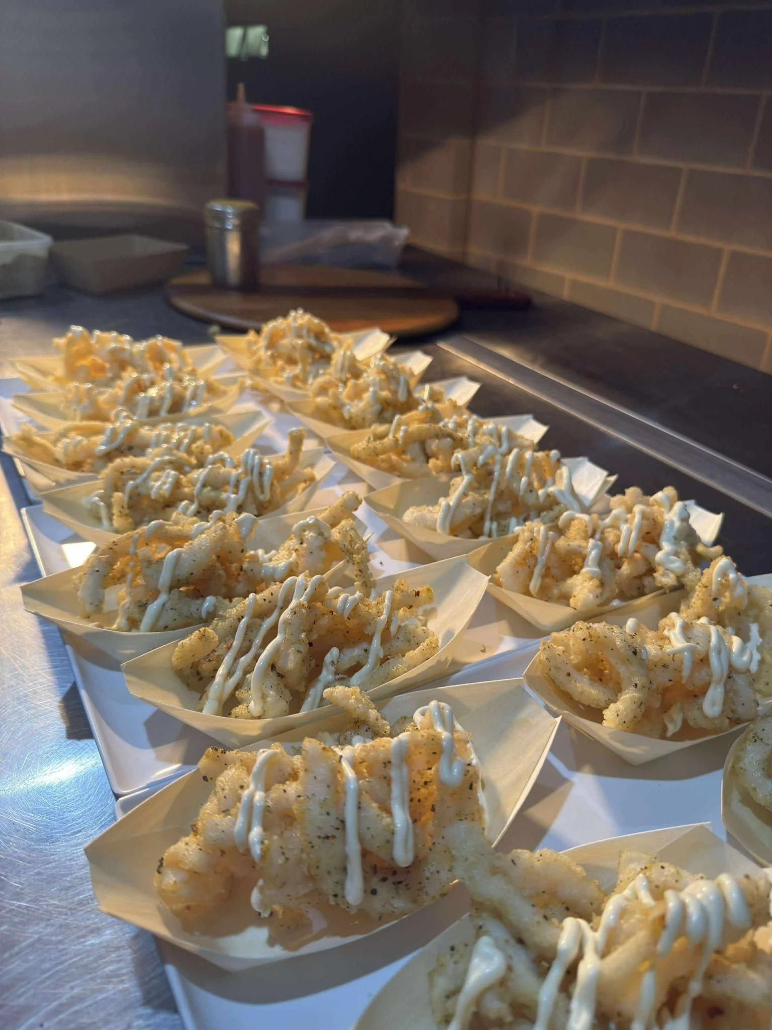 Fried calamari topped with mayonnaise on small paper boats, arranged on a tray in a kitchen setting.