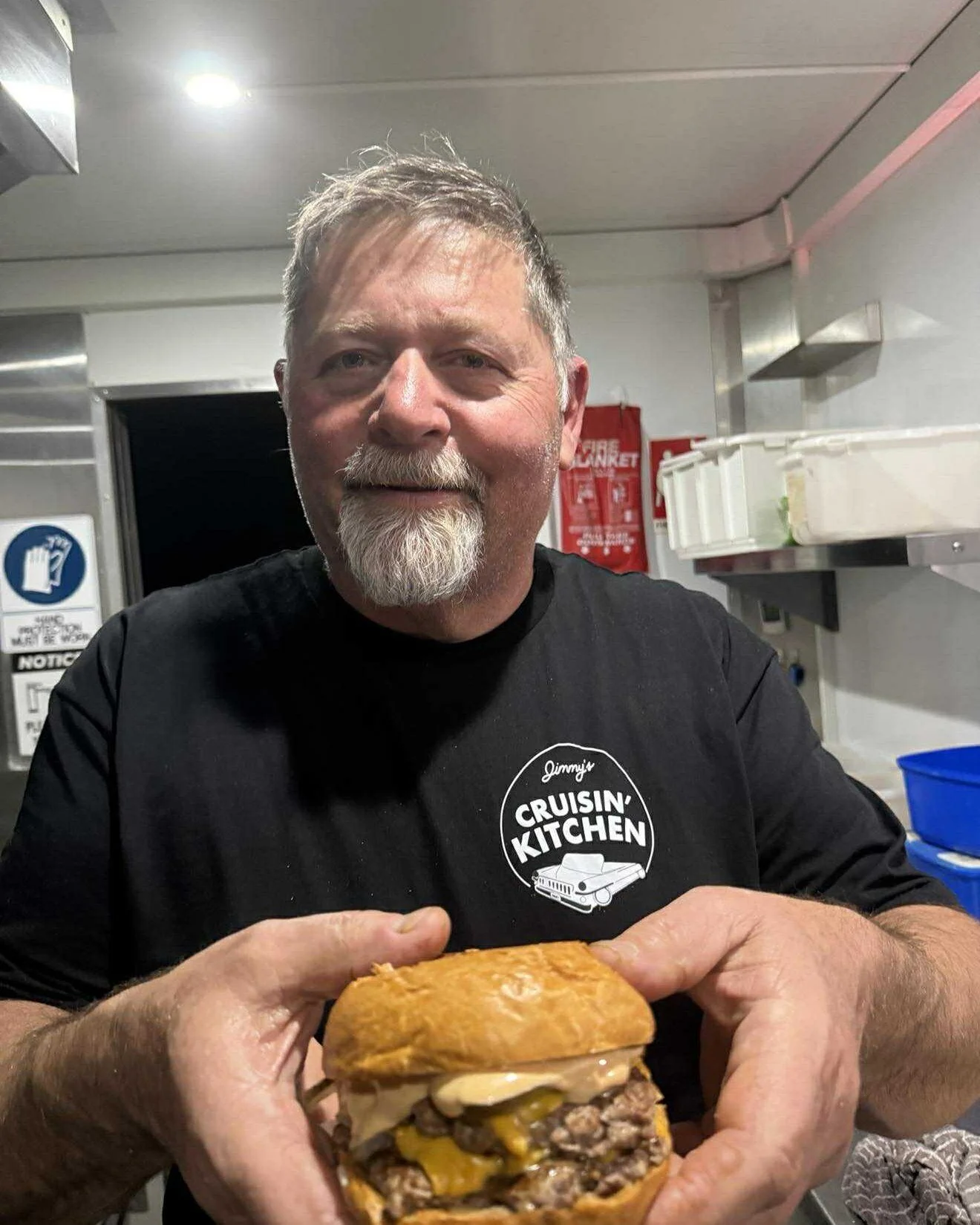 A man with a beard and mustache wearing a black shirt with 'Jimmy's Cruisin' Kitchen' logo, holding a cheeseburger with multiple patties and cheese.