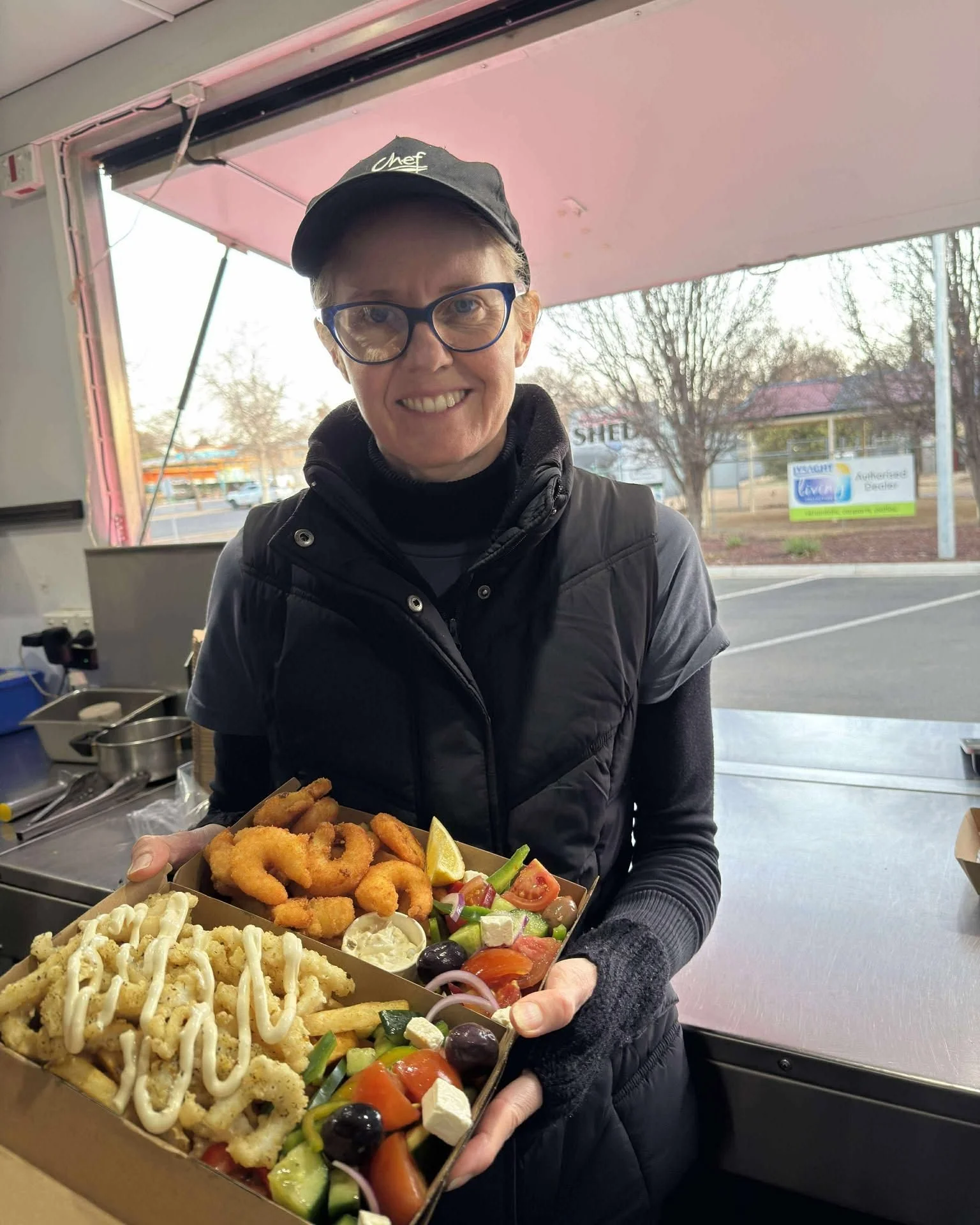 A smiling woman with glasses and a black cap holding two food trays filled with fried calamari, onion rings, and a vegetable salad inside a food stand or truck.