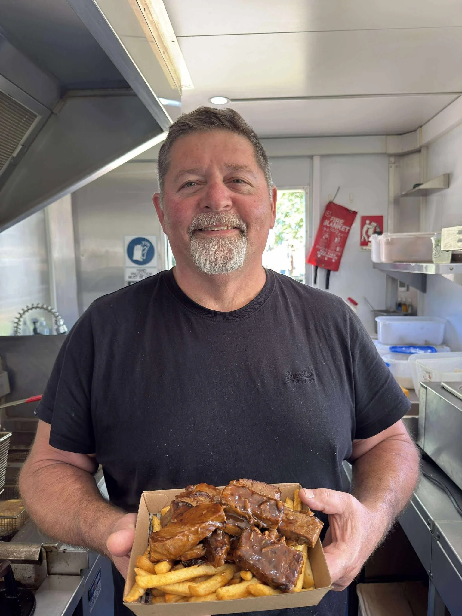 A man with gray hair and beard smiling while holding a box of French fries topped with barbecue ribs in a kitchen or food truck setting.