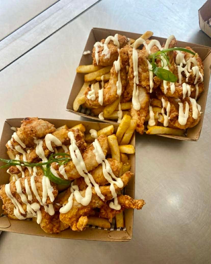 Two trays of fried chicken tenders topped with mayonnaise drizzle served with French fries, garnished with green herbs on a gray table.