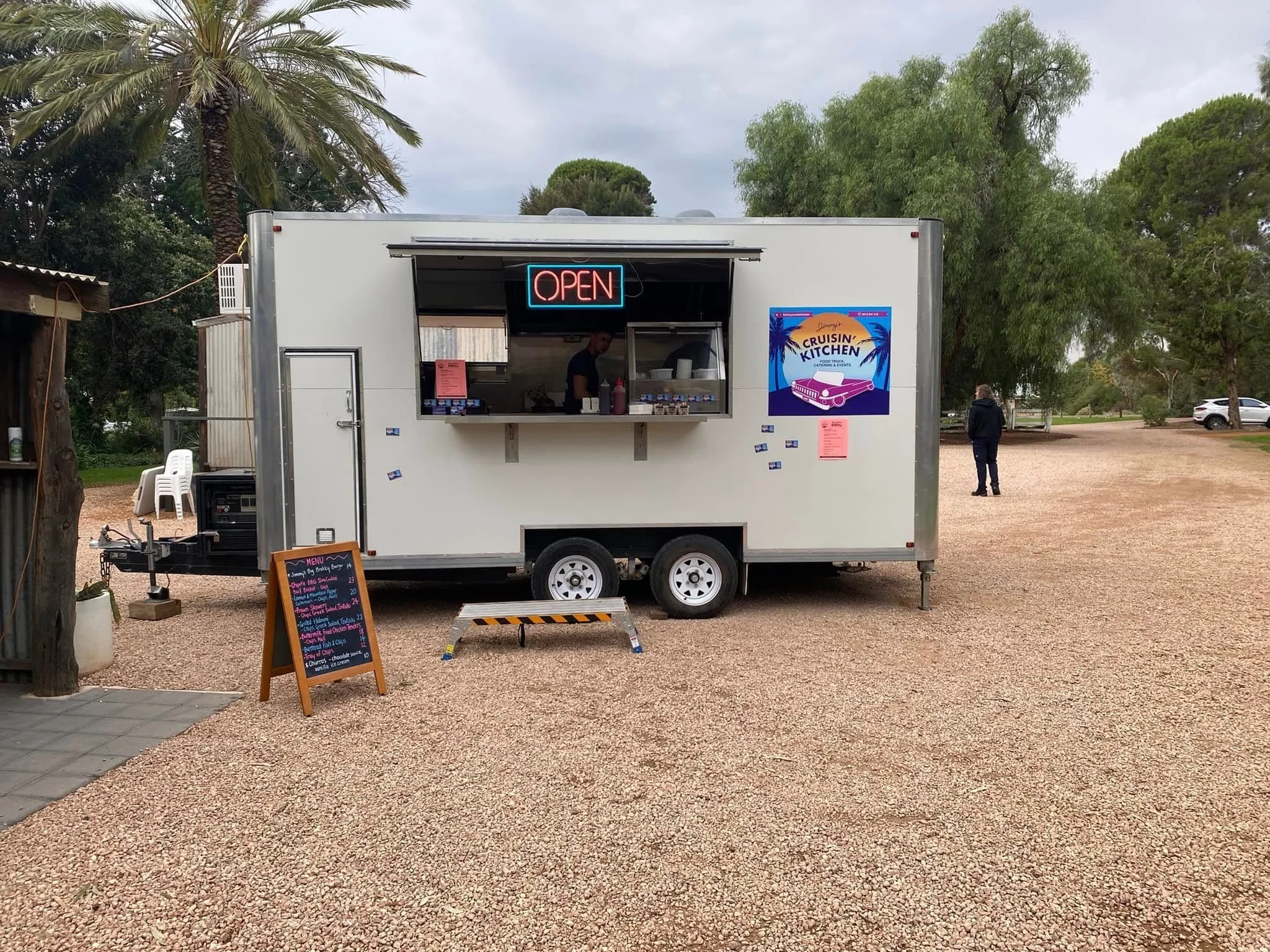 Food truck with an open window, neon 'Open' sign, menu board outside, and a poster on the side, parked on gravel with trees and a cloudy sky in the background.
