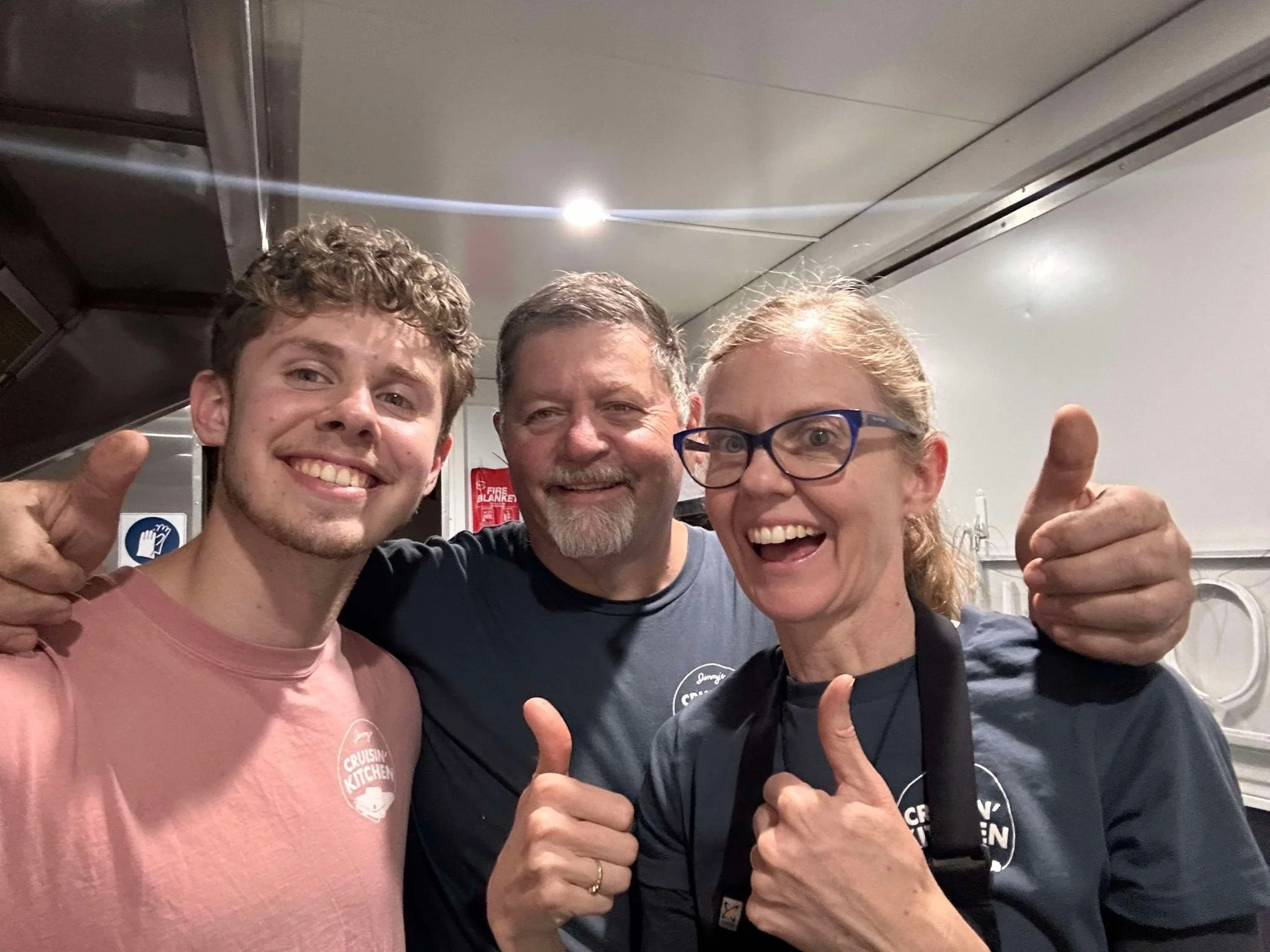 Three smiling people posing together indoors, giving thumbs up and wearing casual clothes.