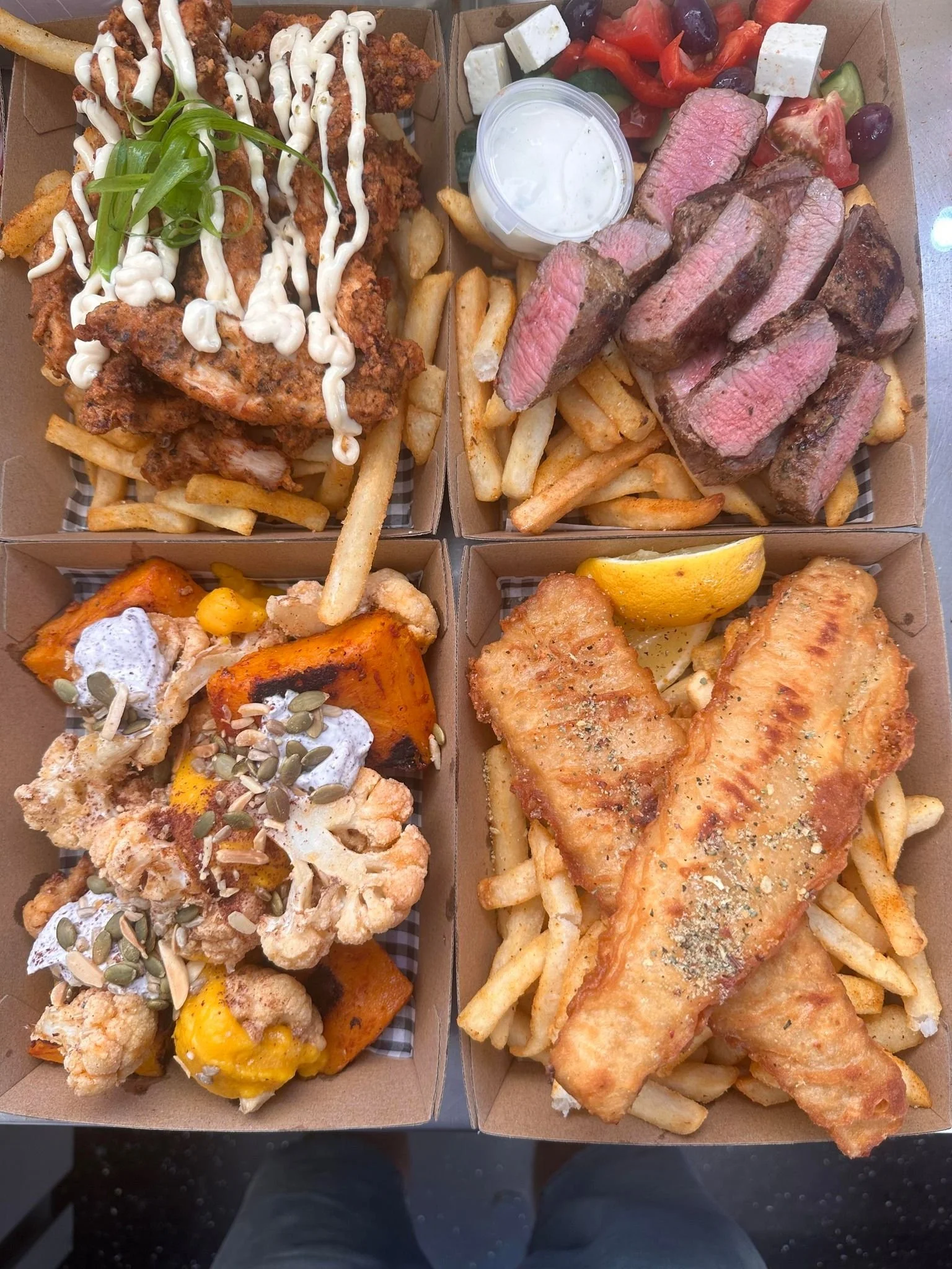 Four food boxes containing fried chicken with slaw, grilled steak with fries, roasted cauliflower with pumpkin and seeds, and fried fish with French fries and lemon wedge.