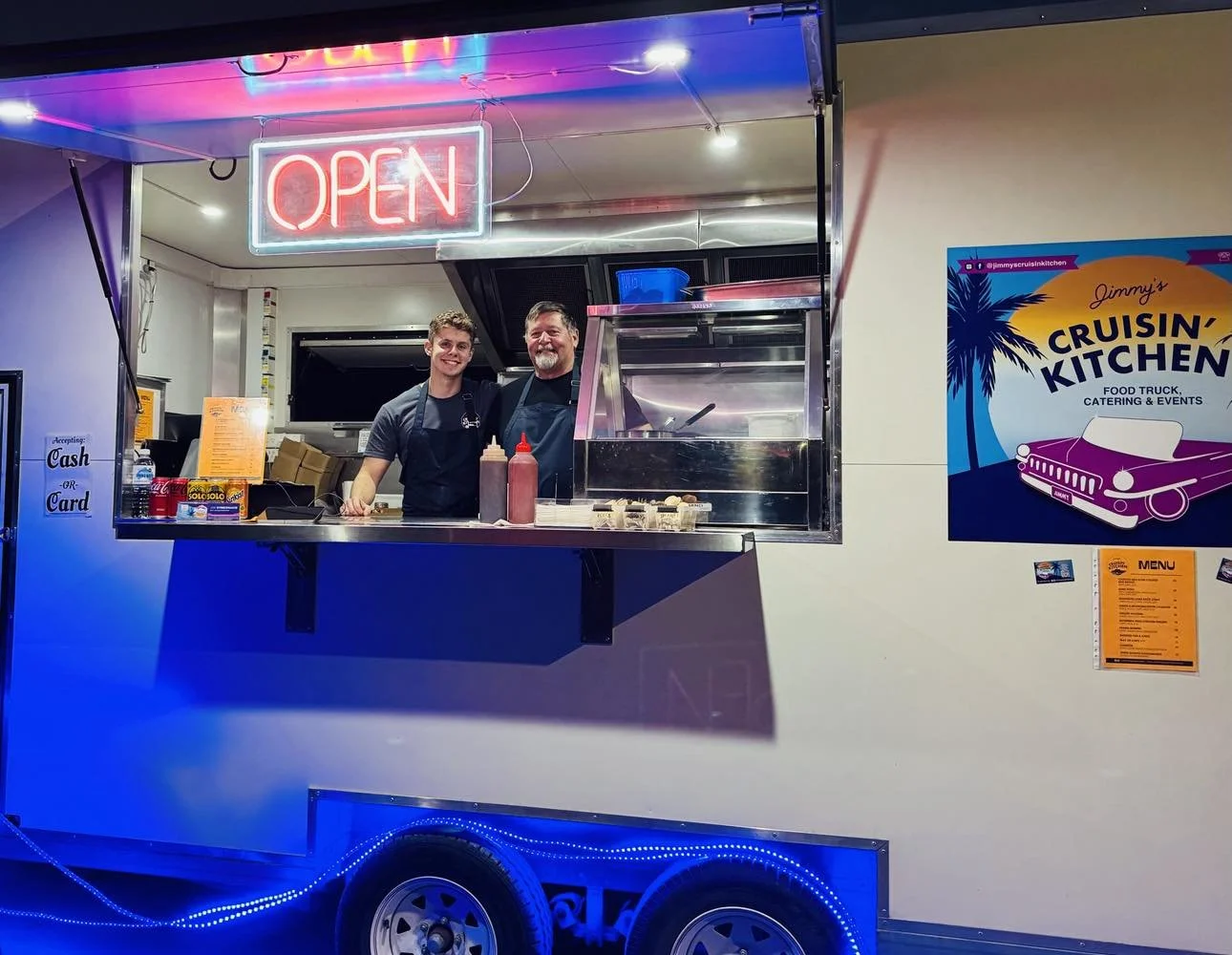Two men in aprons inside a food truck with a neon 'OPEN' sign, colorful neon lights, condiments on the counter, and a 'Jimmy's Cruisin' Kitchen' sign on the wall.