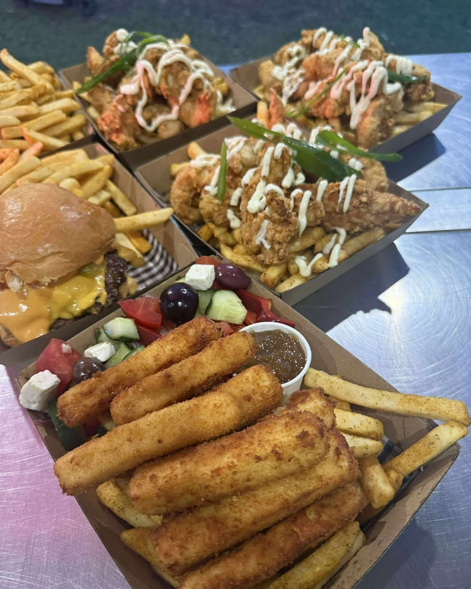 A tray of fried fish sticks with French fries, accompanied by a salad with tomatoes, cucumbers, black olives, feta cheese, and a cup of dipping sauce. In the background, there is a cheeseburger with fries and a dish of fried chicken topped with mayon