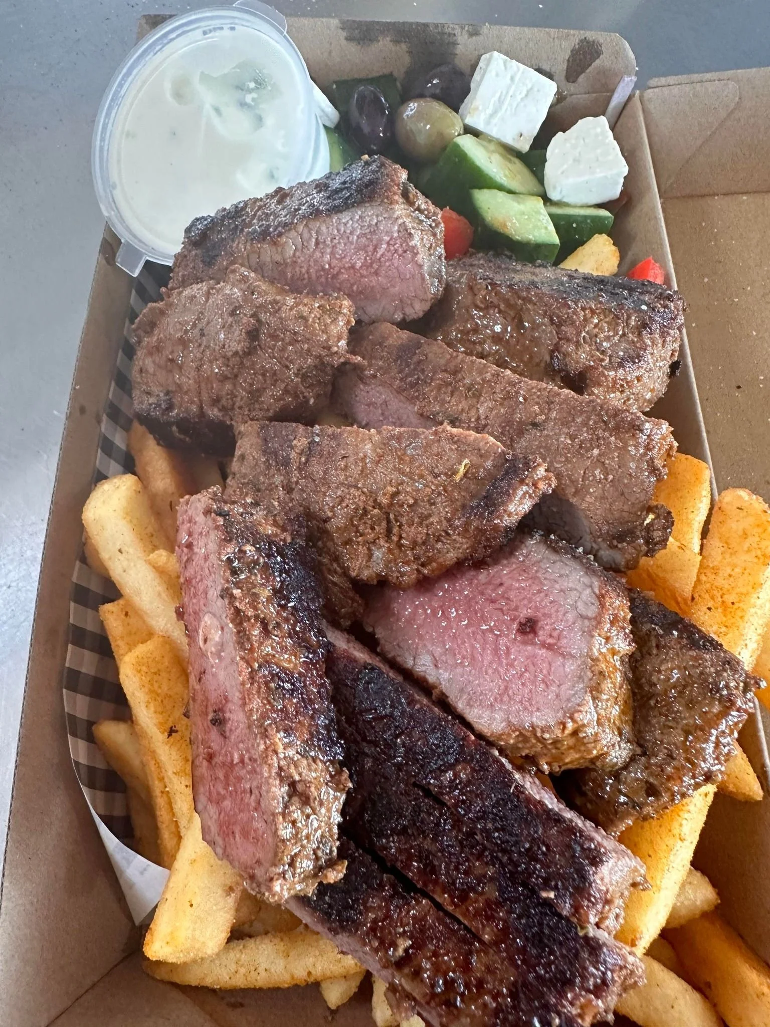 A box of French fries topped with grilled steak, a side of salad with cucumber, olives, and feta cheese, and a small container of dipping sauce.