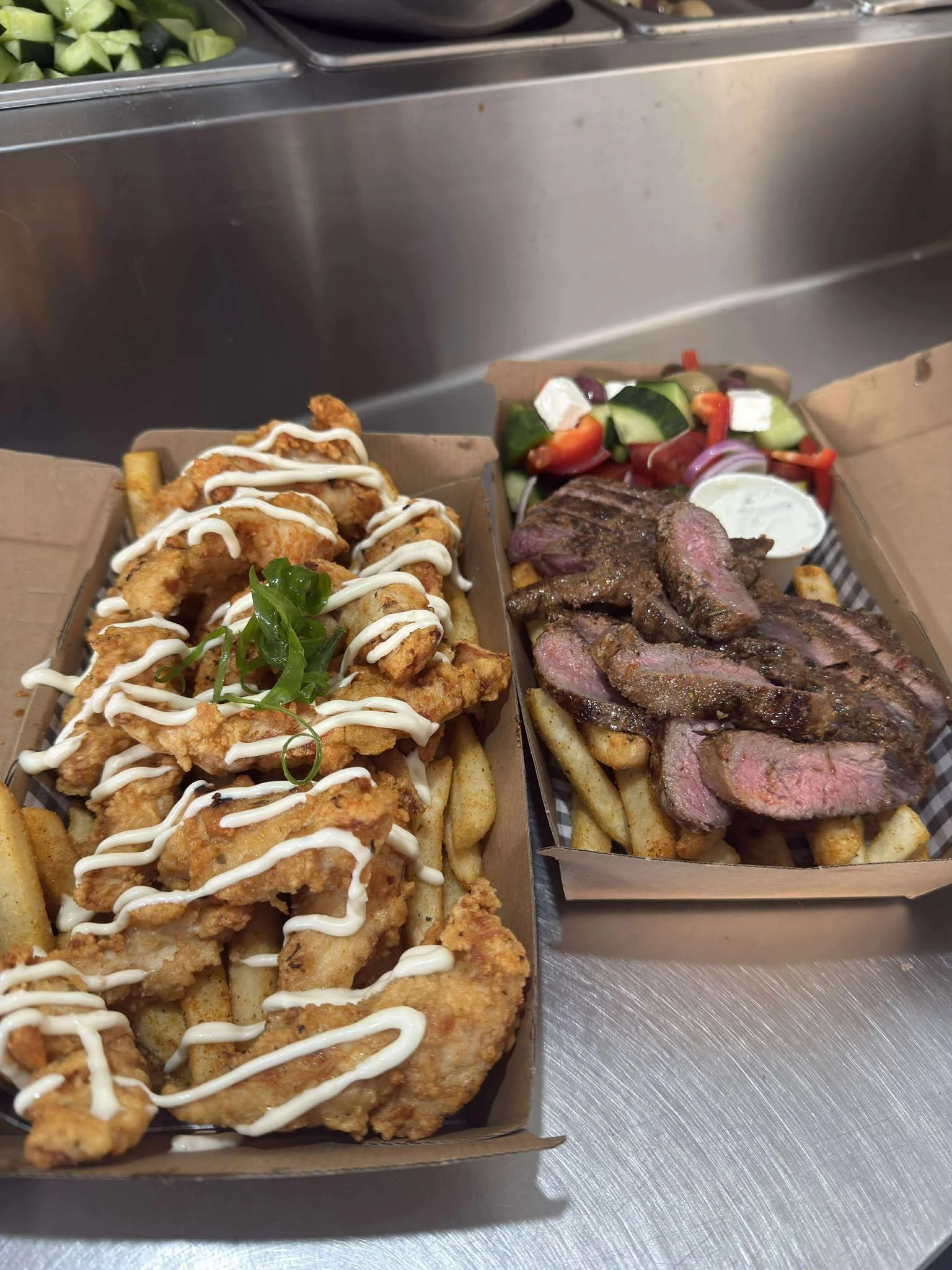 Two food trays; one with fried chicken tenders drizzled with white sauce and garnished with green herbs, served over French fries, and the other with sliced grilled steak, French fries, and a side salad of cucumbers, tomatoes, red onions, feta cheese