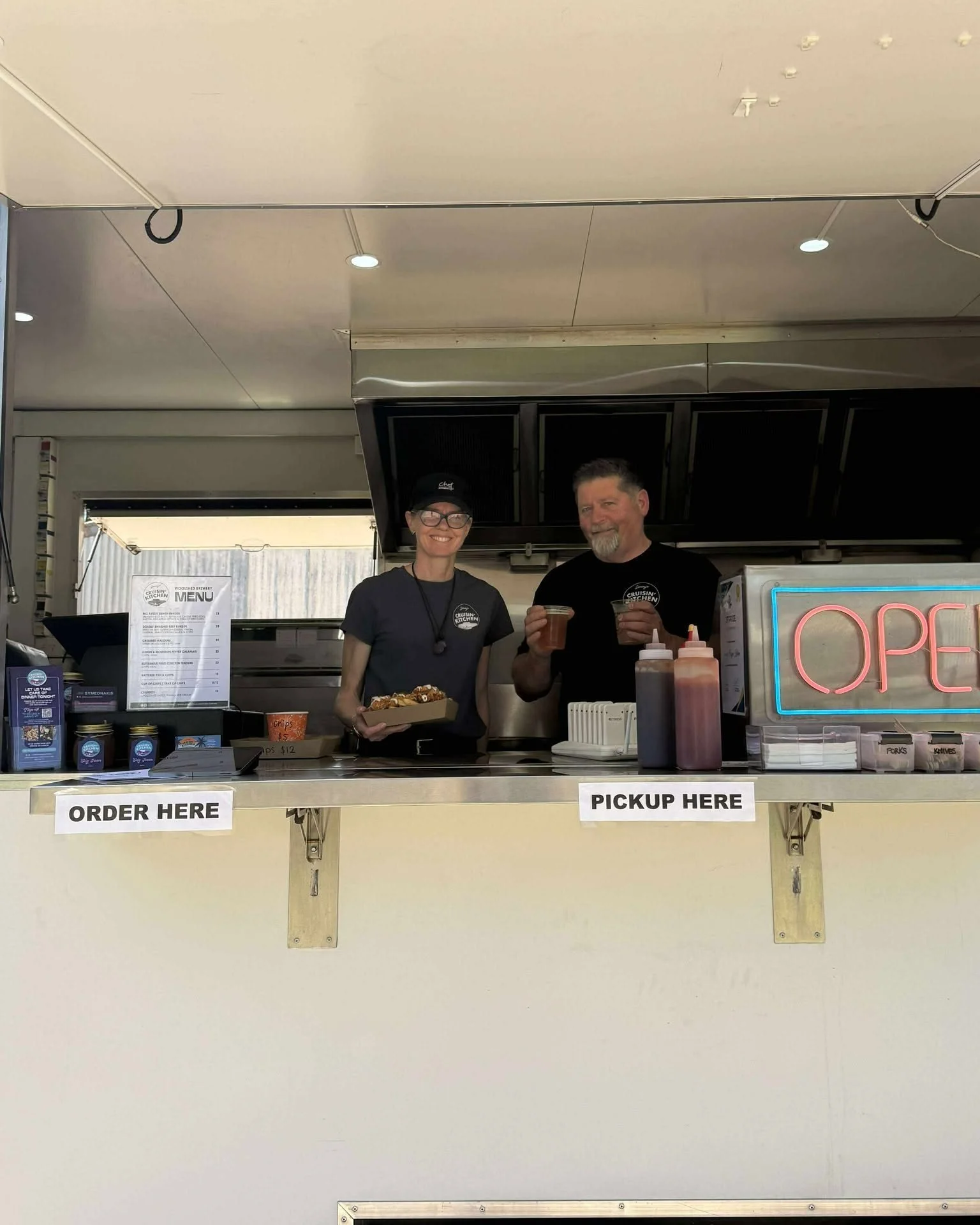Two smiling food vendors standing behind a counter with a neon 'OPEN' sign, condiments, and a menu, serving food and drinks in a food stall.