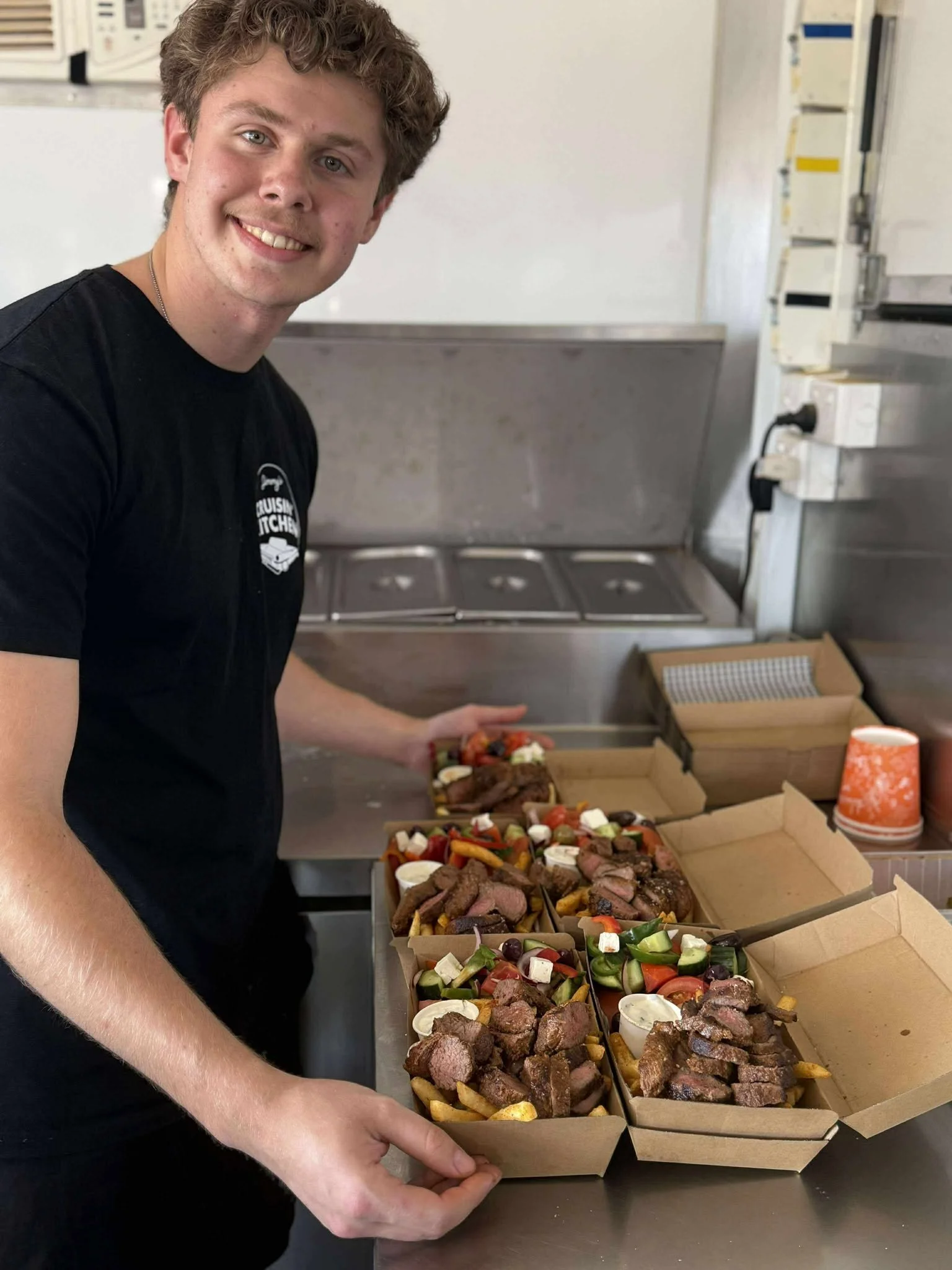 A smiling young man in a black t-shirt holding a box of mixed grilled meats and vegetables, standing in a kitchen with food containers in the background.