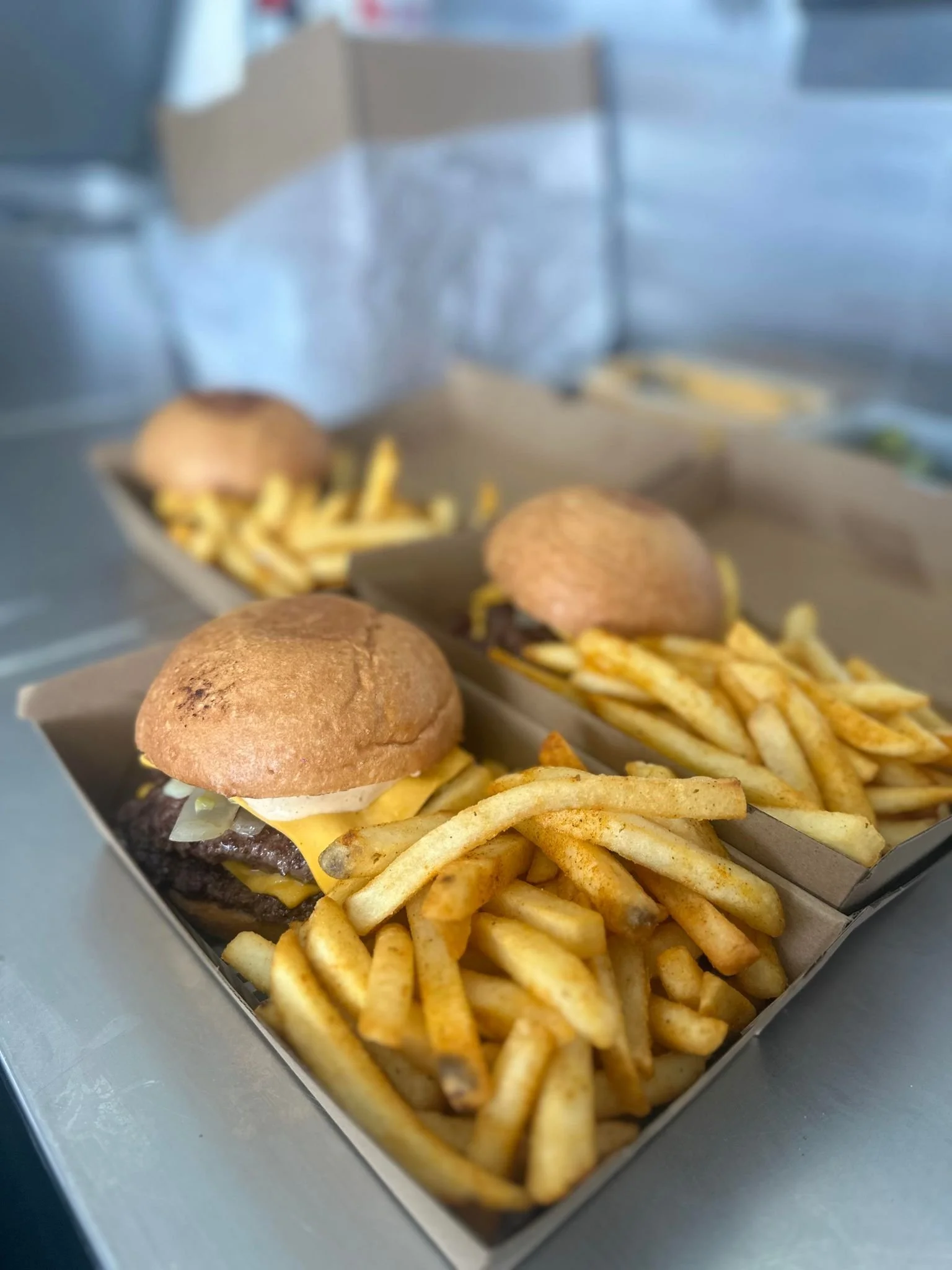 Three trays of cheeseburgers with French fries on a stainless steel surface.