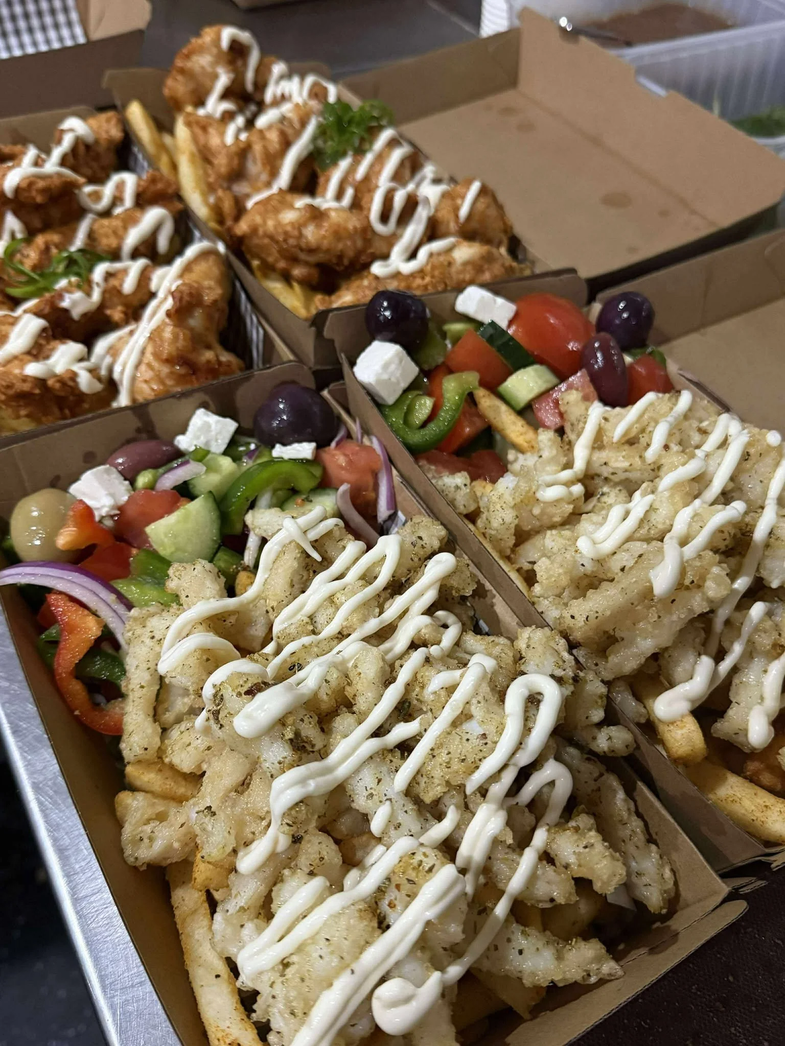 Three takeout boxes filled with fried fish topped with mayonnaise sauce, and a side of fresh vegetable salad with tomatoes, cucumbers, olives, red onions, and feta cheese, with fried French fries in one of the boxes.