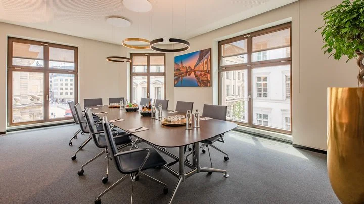 Empty modern conference room with a large oval table, black chairs, large windows, a colorful cityscape artwork, and decorative ceiling lights.