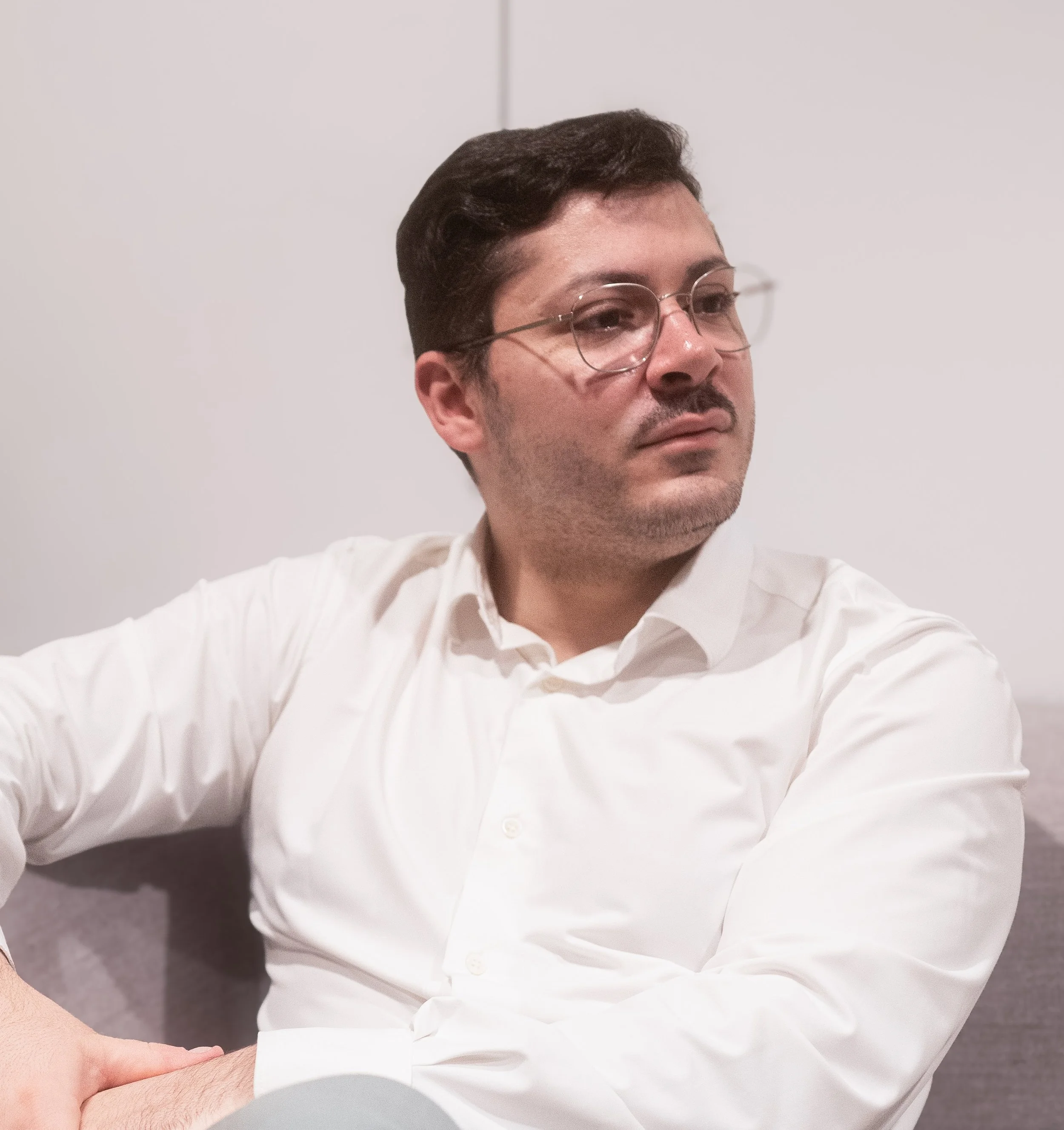 A man with dark brown hair, wearing glasses and a white shirt, sitting on a gray couch, looking to his right with a neutral expression.