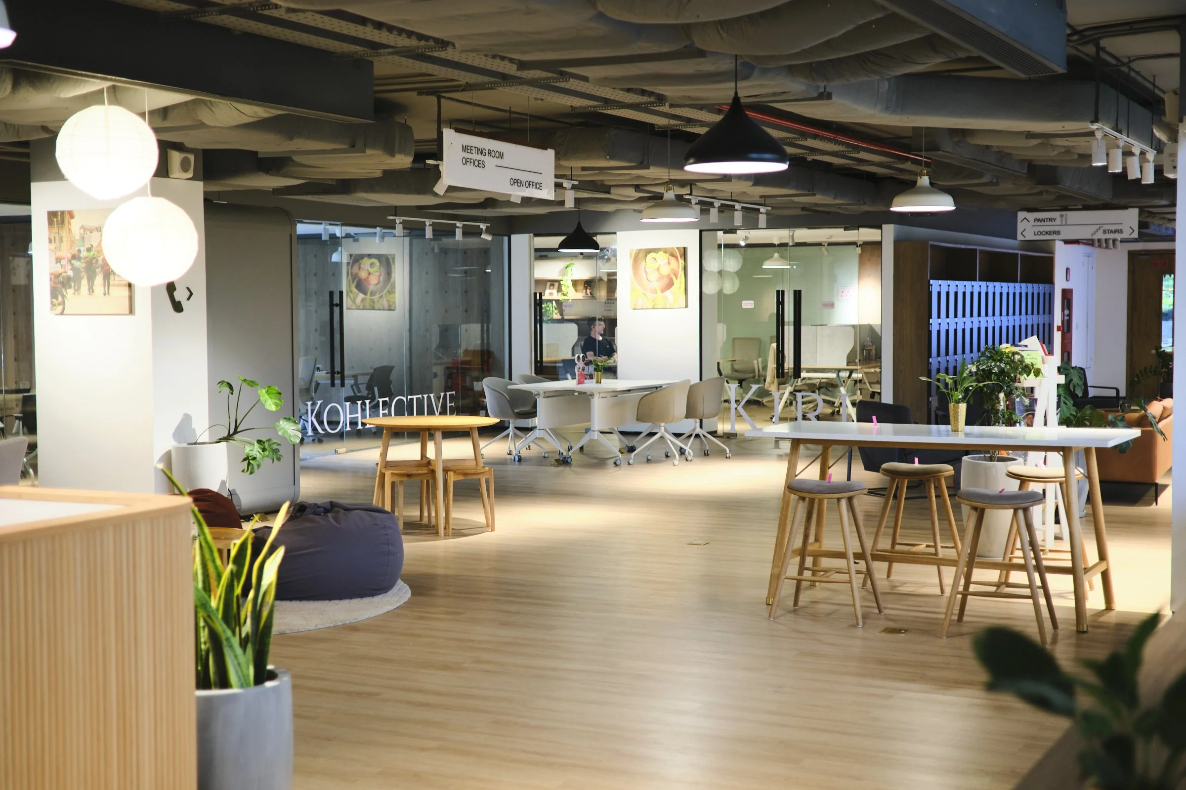 Modern office space with wooden floors, several tables and chairs, green plants, glass meeting rooms, and signs indicating locations such as meeting rooms, offices, open office, pantry, lockers, and stairs.