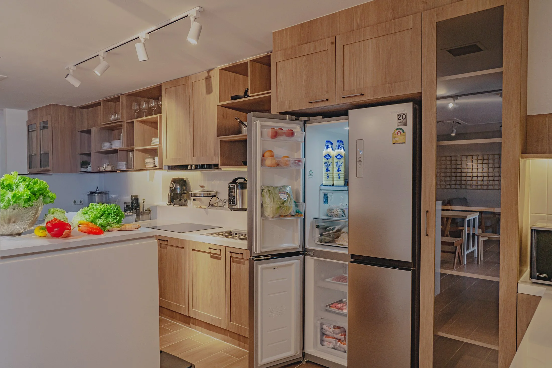 Modern kitchen with wooden cabinets, open refrigerator with food and drinks, and a countertop with vegetables and kitchen appliances.