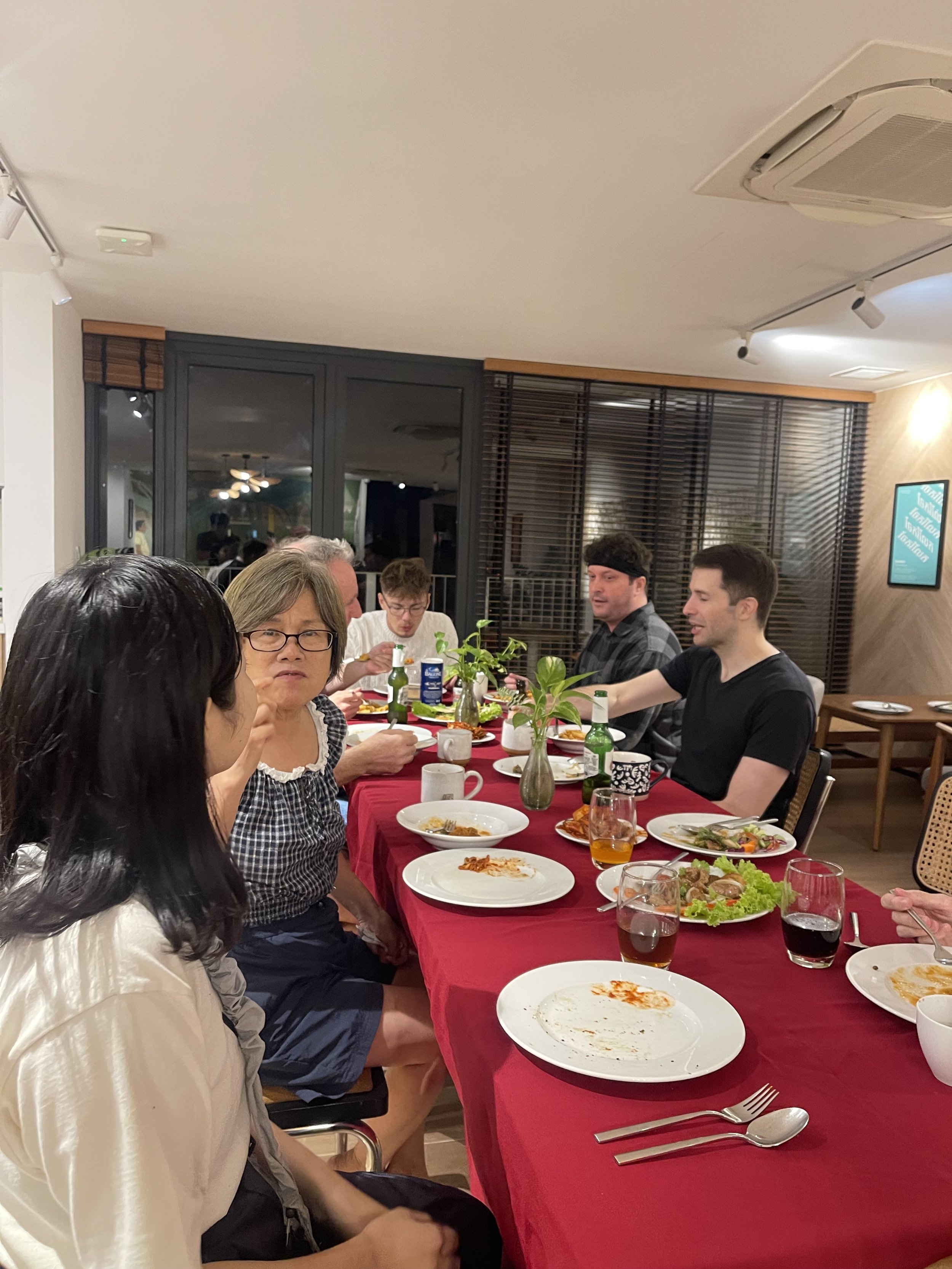 People sitting at a dining table after a meal in a restaurant. Some plates have leftover food, and there are drinks, utensils, and a small plant centerpiece. The background shows large windows with blinds and a flat-screen TV on the wall.