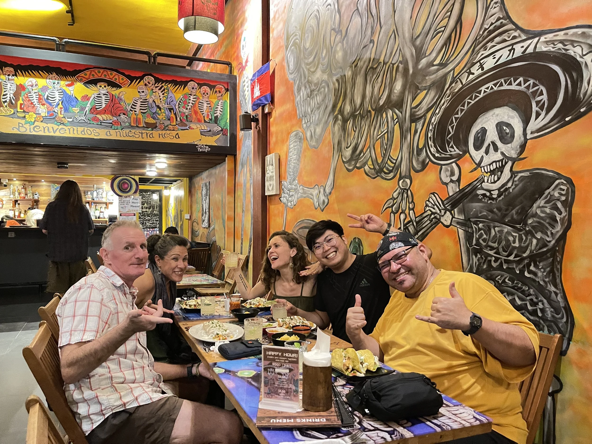 Group of smiling people at a restaurant table with plates of food, drinks, and utensils, with colorful Mexican-themed mural art and decorations in the background.