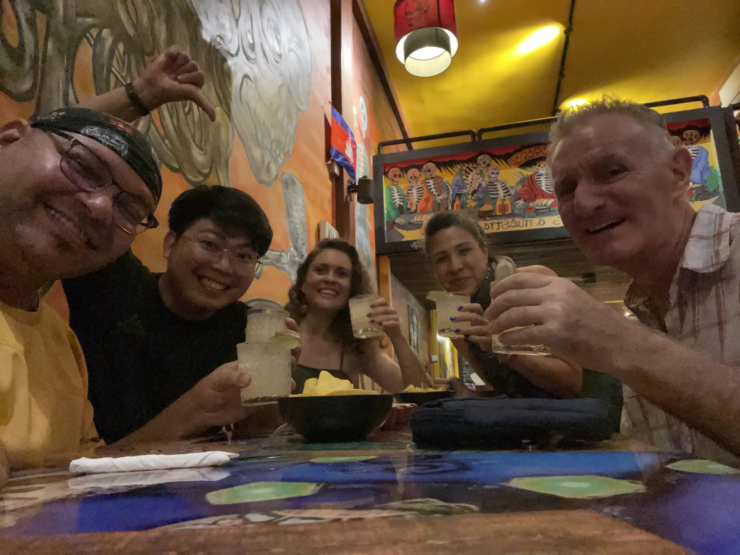A group of people sitting at a table in a restaurant, holding and enjoying drinks, with vibrant, colorful wall art and decorations in the background.