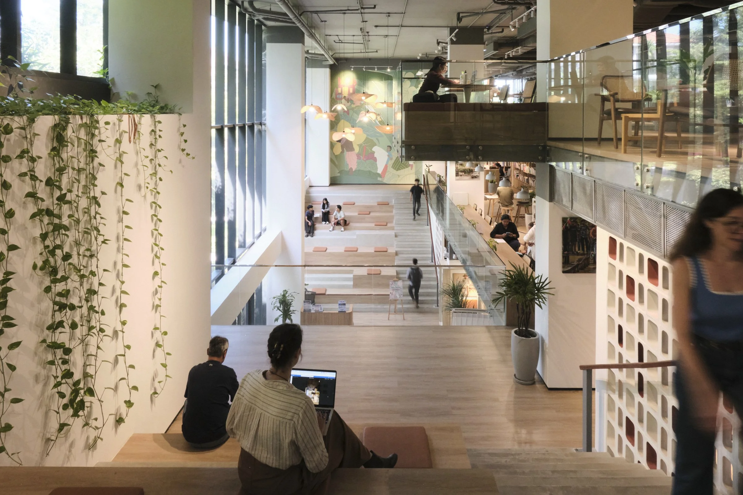 Interior of a modern, multi-story building with people working, sitting, and walking in different areas, including a person seated on a ledge using a laptop and others sitting on stairs and at tables, with large windows letting in natural light and d