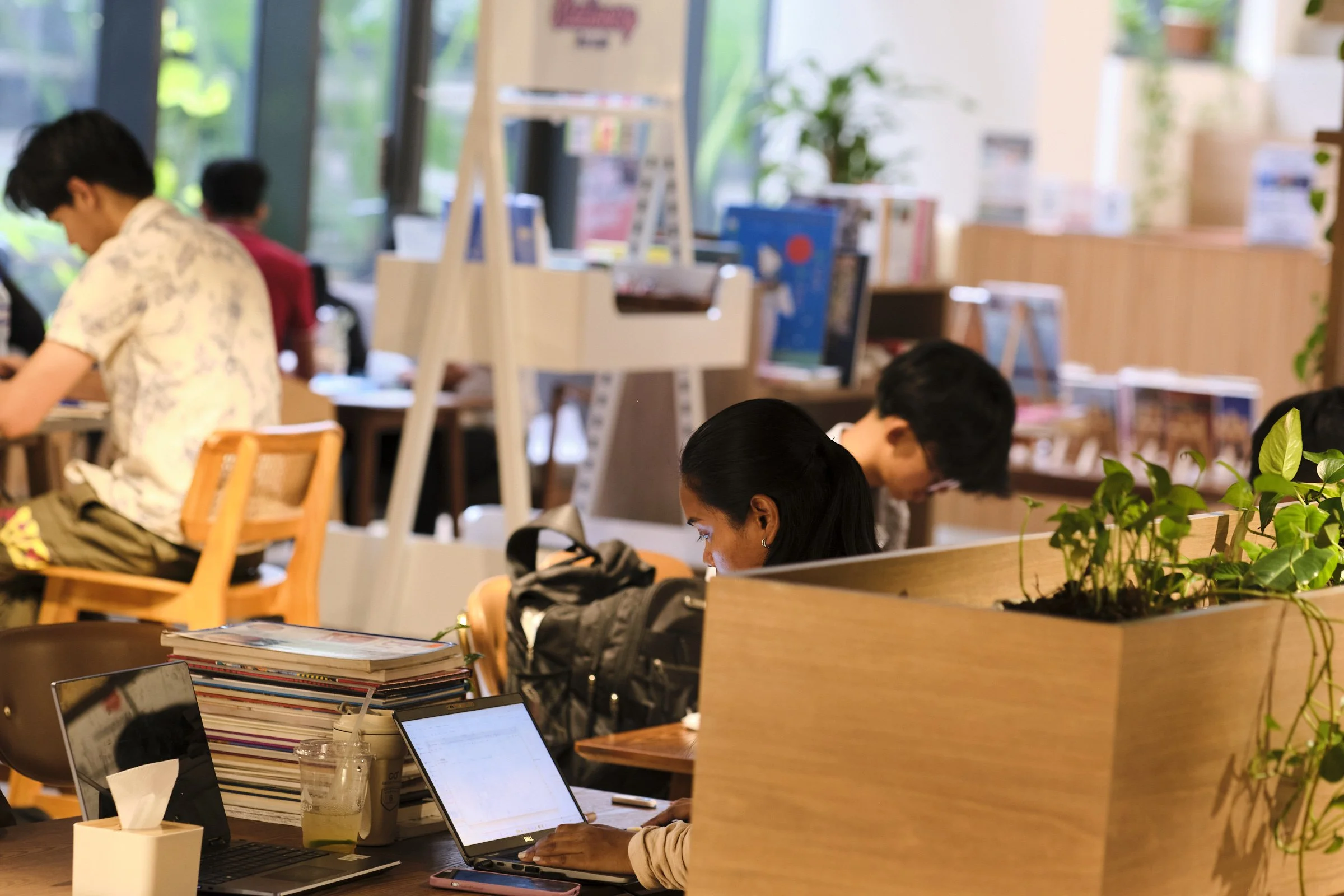 People working on laptops at a co-working space or cafe, with laptops, notebooks, and plants on the tables, and large windows in the background.