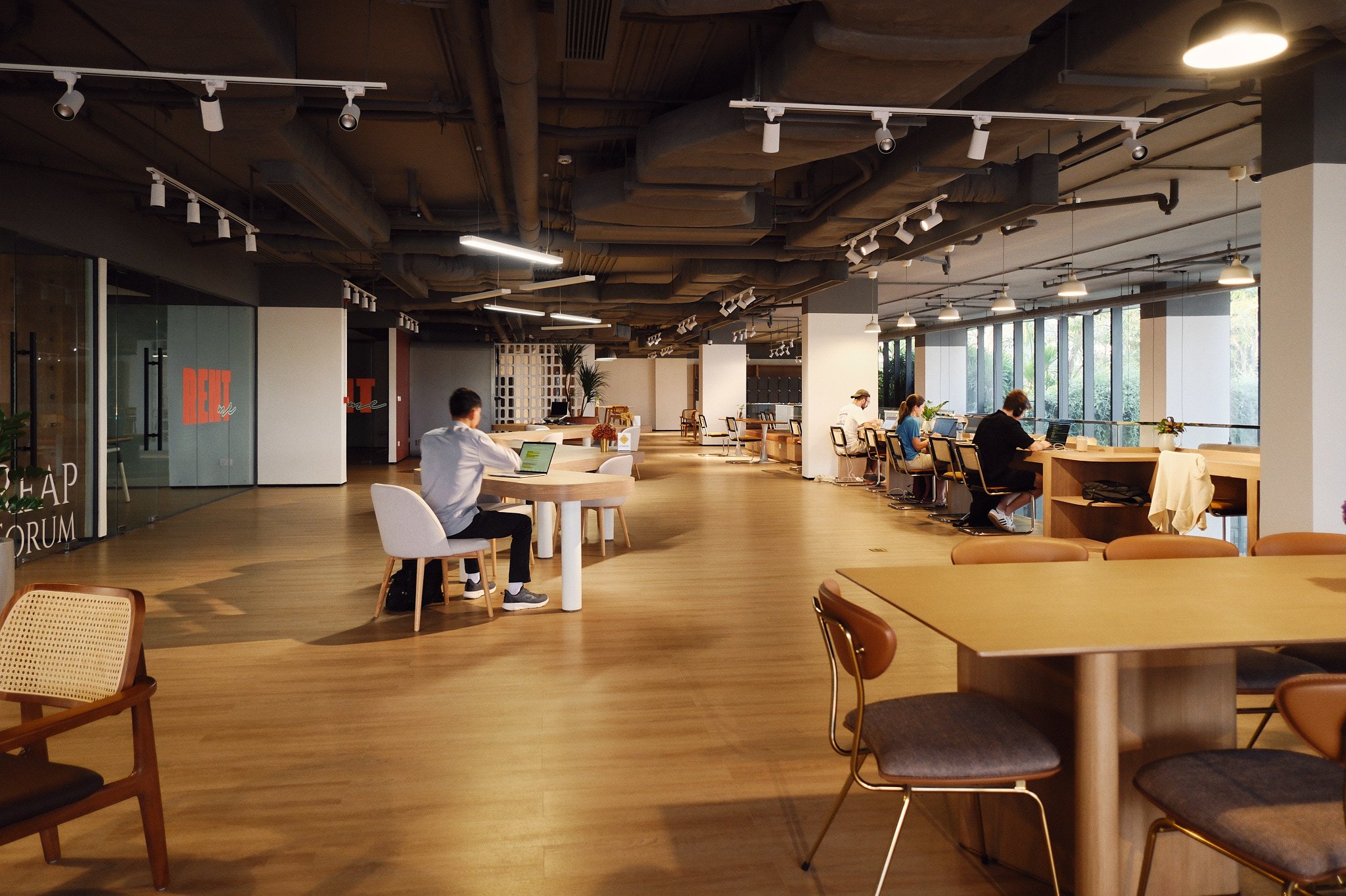 Office space with several people working on laptops, large windows on the right side, modern ceiling with exposed pipes, and a variety of tables and chairs for seating.