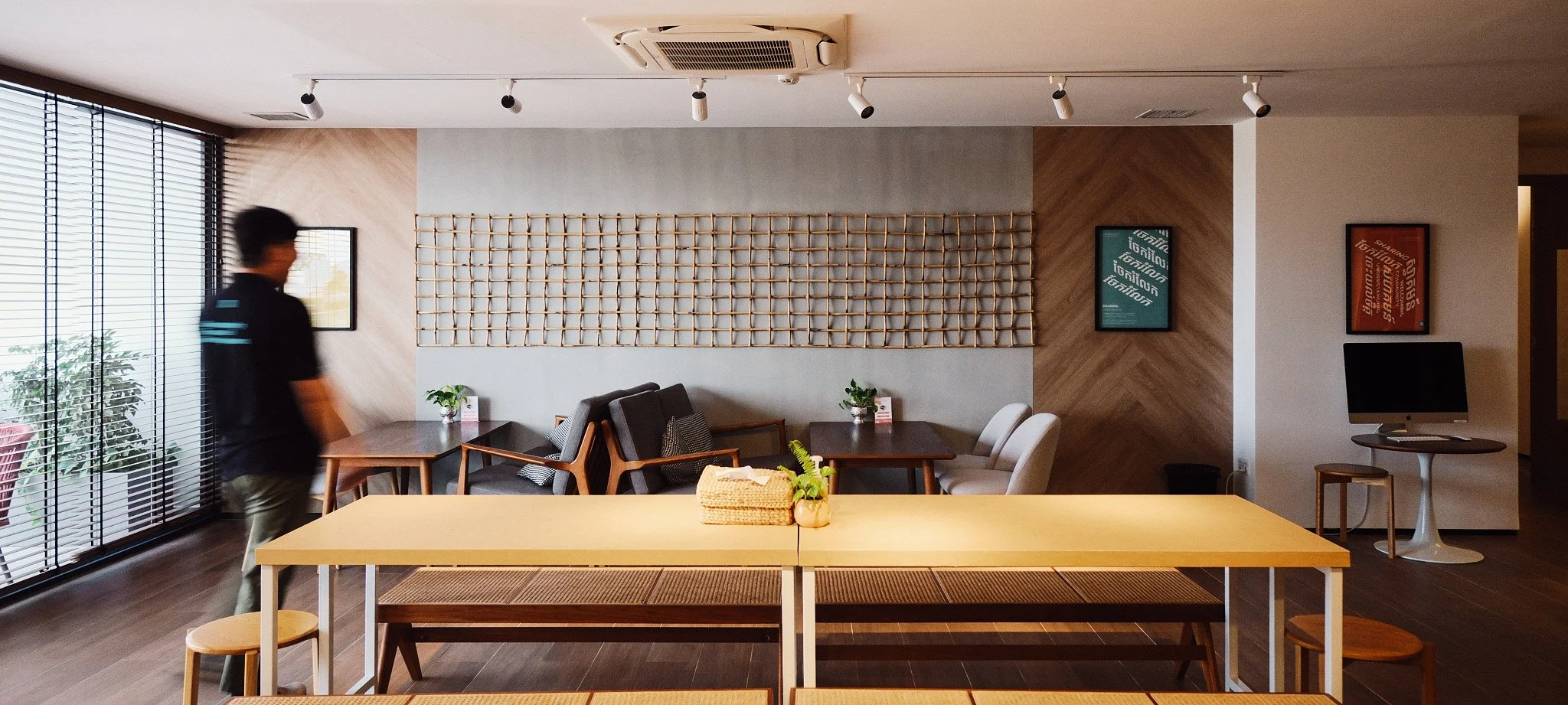 Modern coffee shop interior with several small tables and chairs, a sitting area with armchairs, framed artwork, a computer, and decorative wall hangings, with a person in motion near the window.