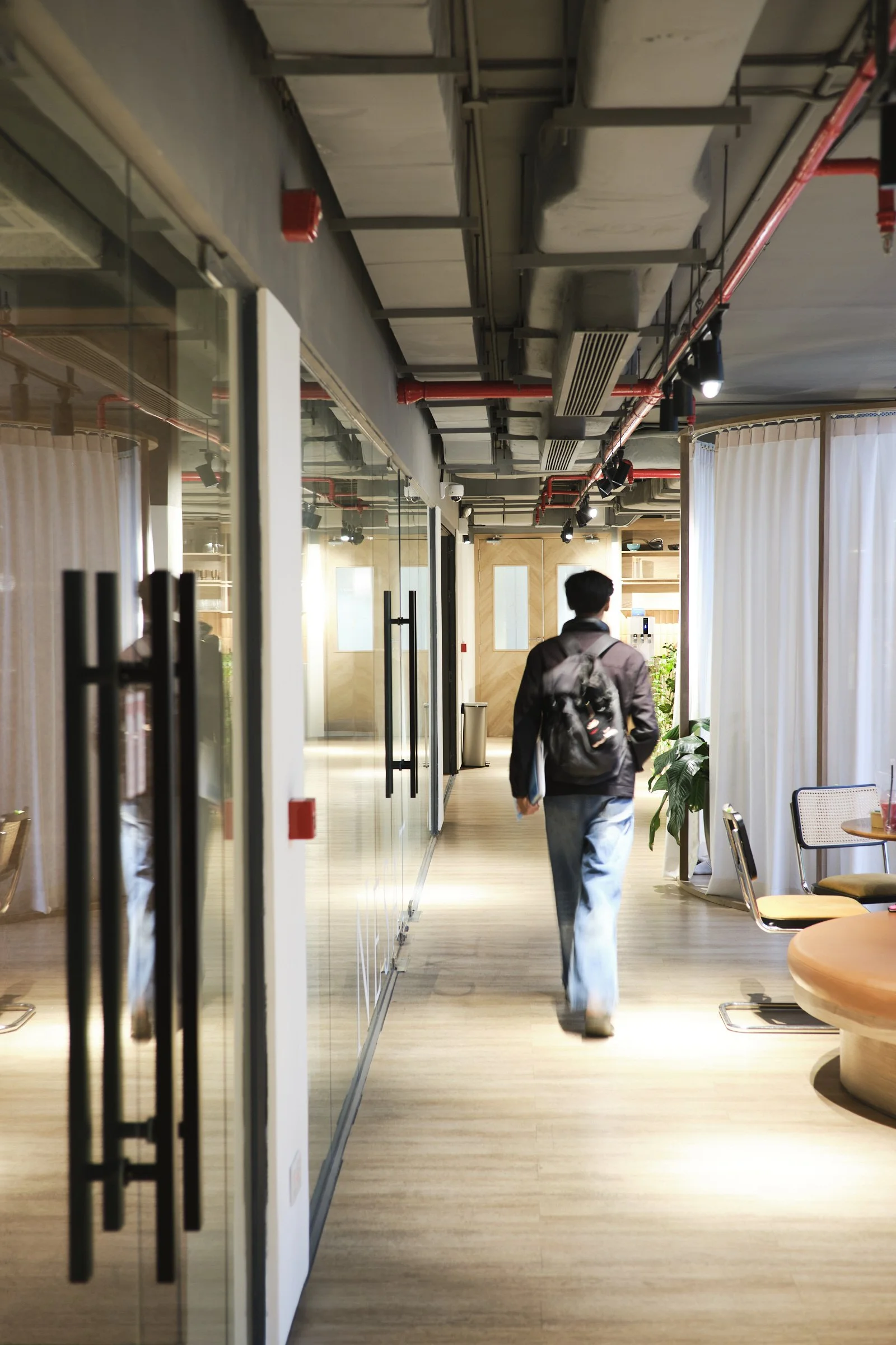 A person walking inside a modern office or coworking space, carrying a backpack. The space features glass partitions, wooden flooring, and natural light coming through white curtains.