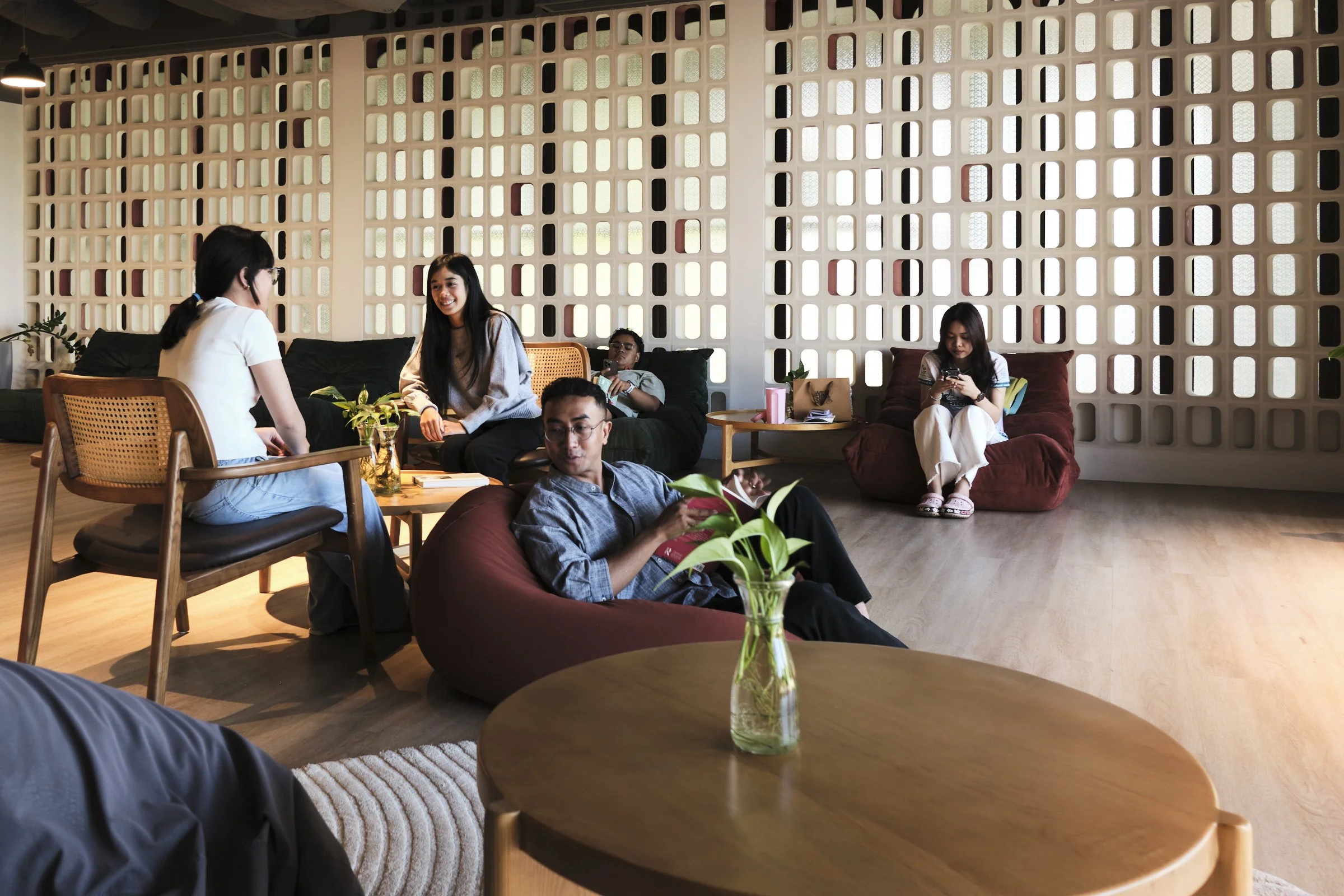 A group of diverse people, including women and men, sitting and socializing in a cozy, modern lounge with a decorative brick wall, plants, and wooden furniture.