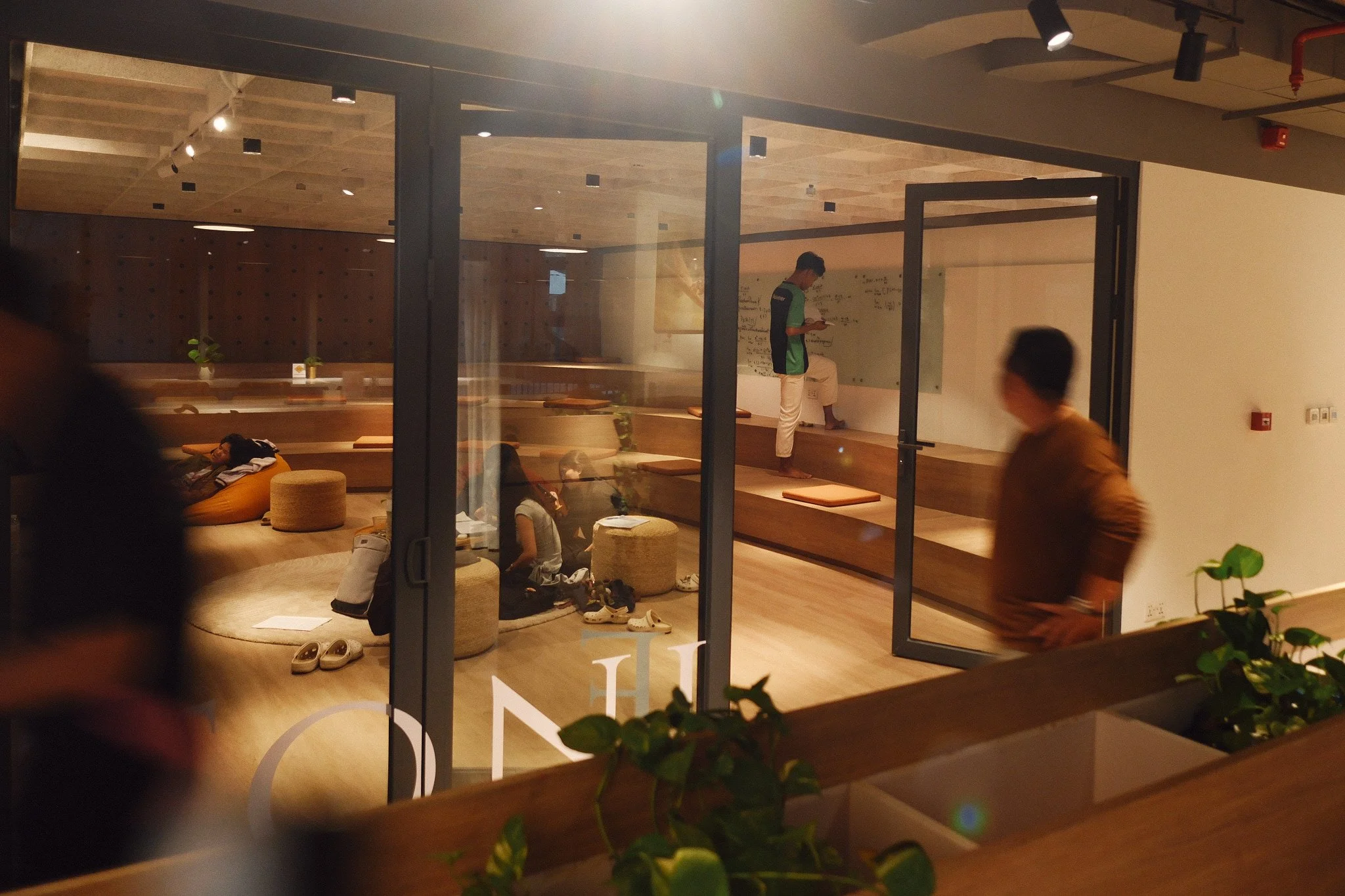 View through a glass window of a modern lounge area with several people, cushions, potted plants, and a whiteboard with writing on the wall.