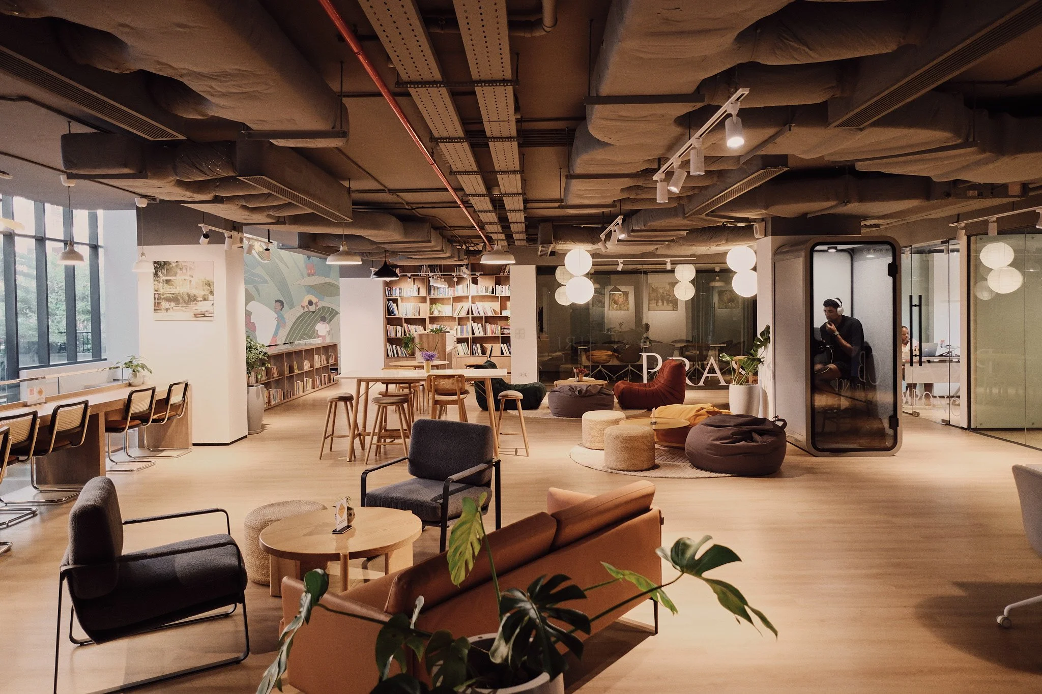 Interior of a modern coworking space or library with various seating arrangements, bookshelves, and decor including plants and artwork, illuminated with hanging lights and natural light from large windows.